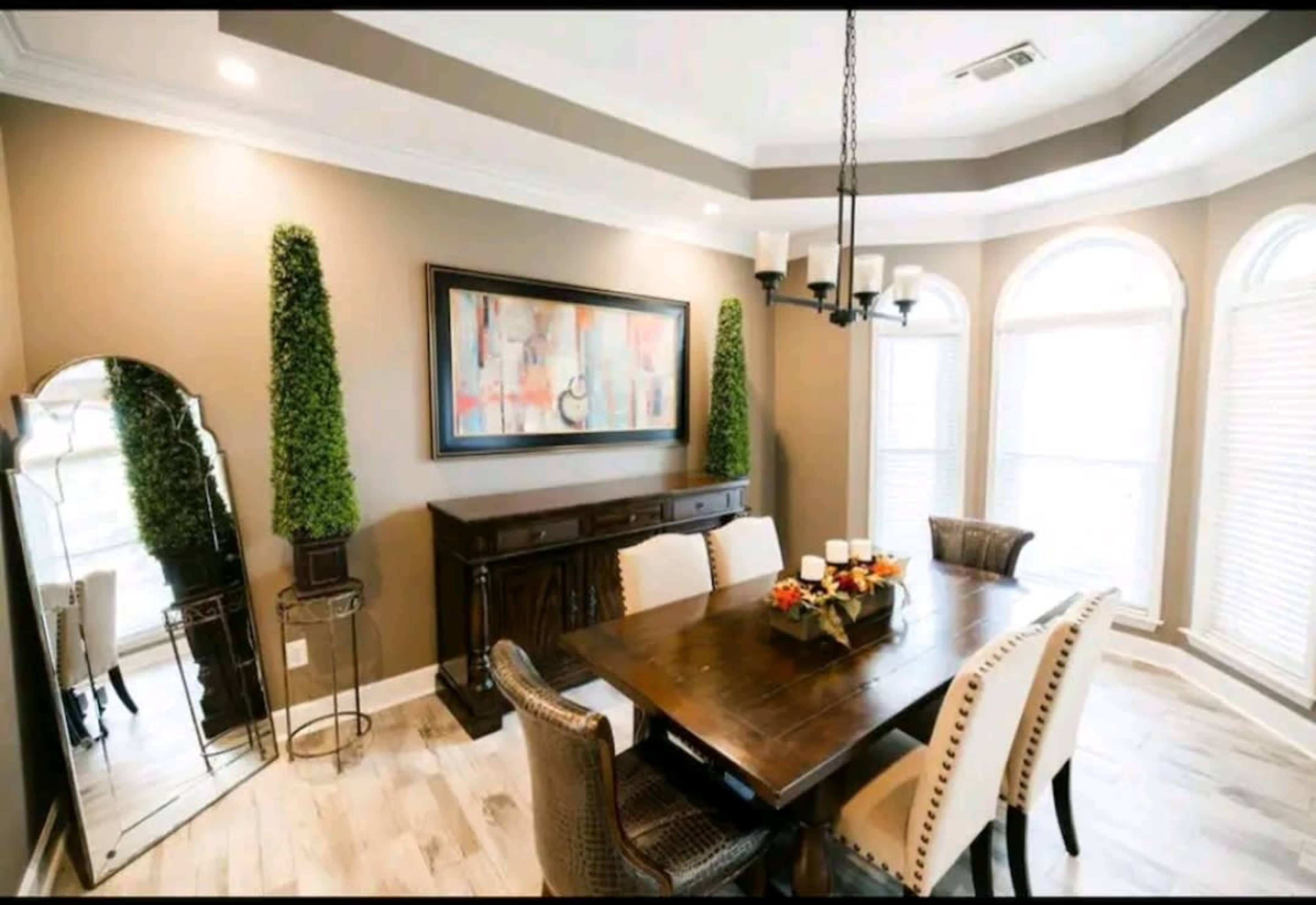 A dining room features a wooden table surrounded by upholstered chairs, with a mirror and decorative greenery against the walls.