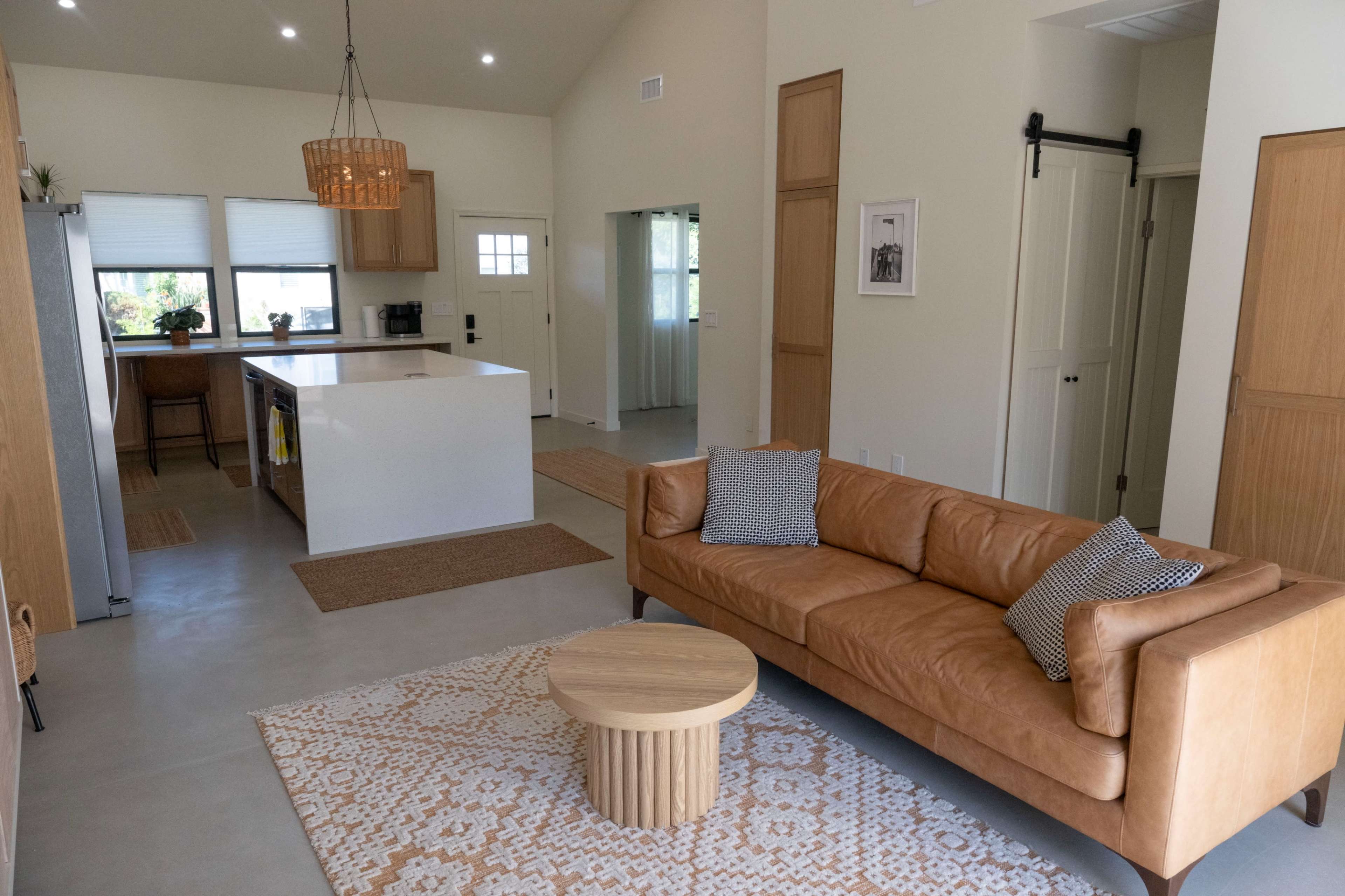 The image shows a modern living room with a tan leather sofa, a round wooden coffee table, and a well-lit open kitchen area in the background.