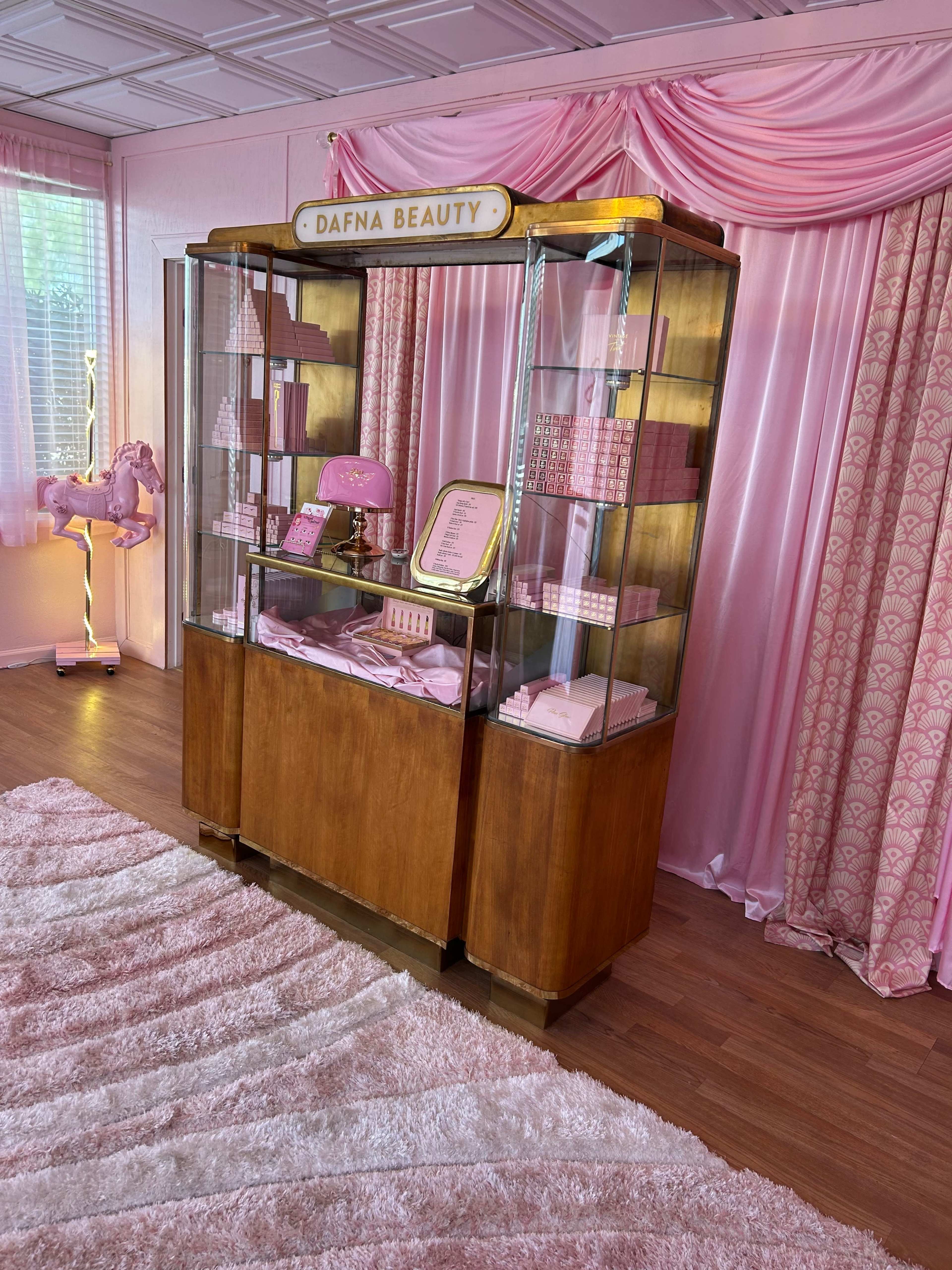 A display cabinet labeled "DAFNA BEAUTY" filled with pink beauty products, set against a backdrop of pink drapes and a plush rug.