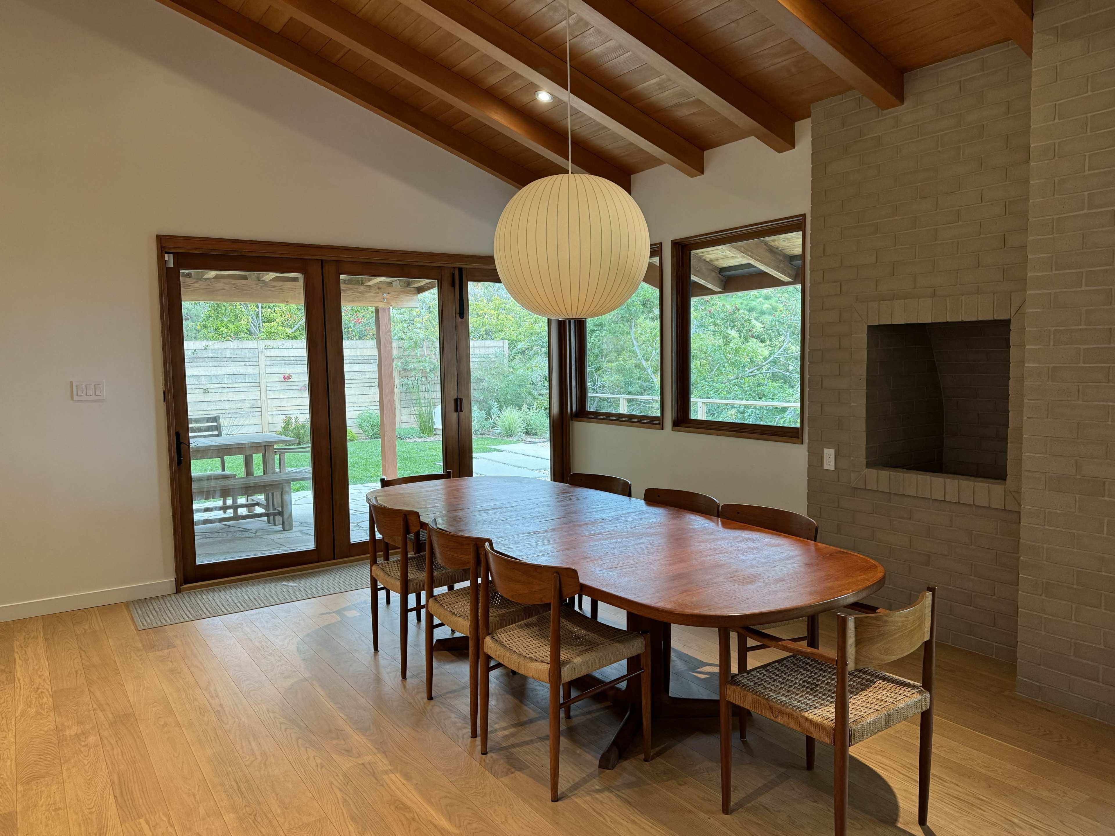 A modern dining area features a wooden table surrounded by chairs, with a large pendant light above, and large windows overlooking a garden.