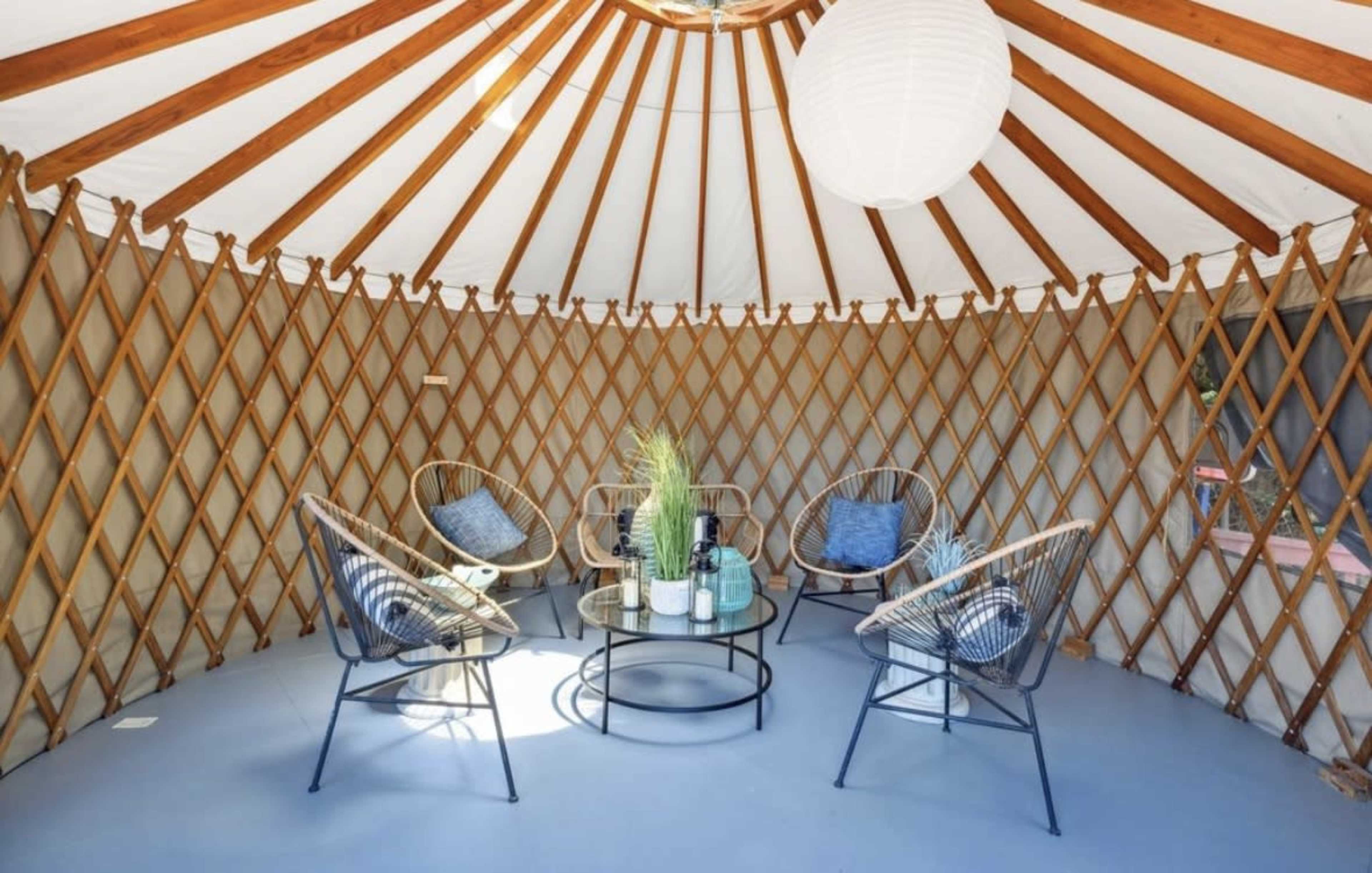 The image shows a circular interior of a yurt furnished with four chairs and a small round table, featuring a lantern and plants.