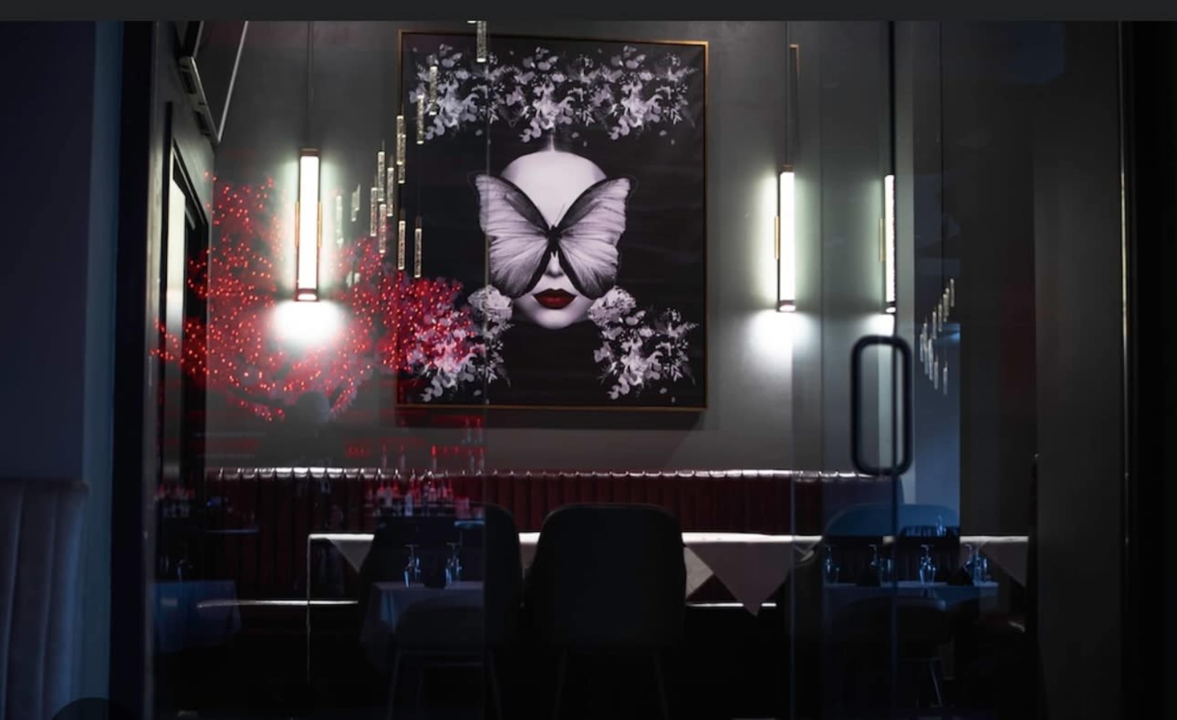 A large artwork featuring a face with a butterfly covering the eyes hangs on the wall of a dimly lit restaurant with tables set for dining.