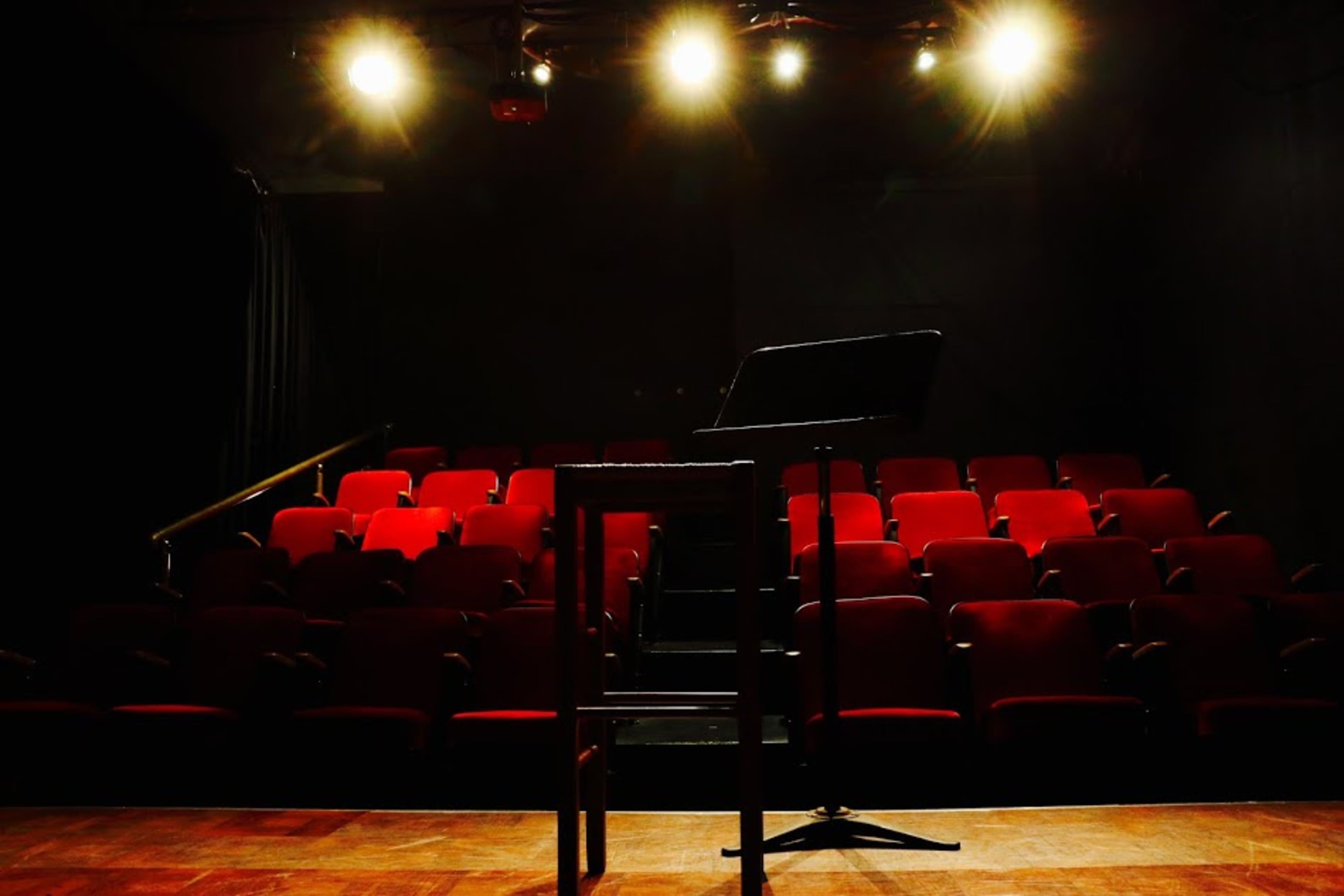 A dimly lit theater stage with a music stand in the foreground and red theater seats arranged in rows behind it.
