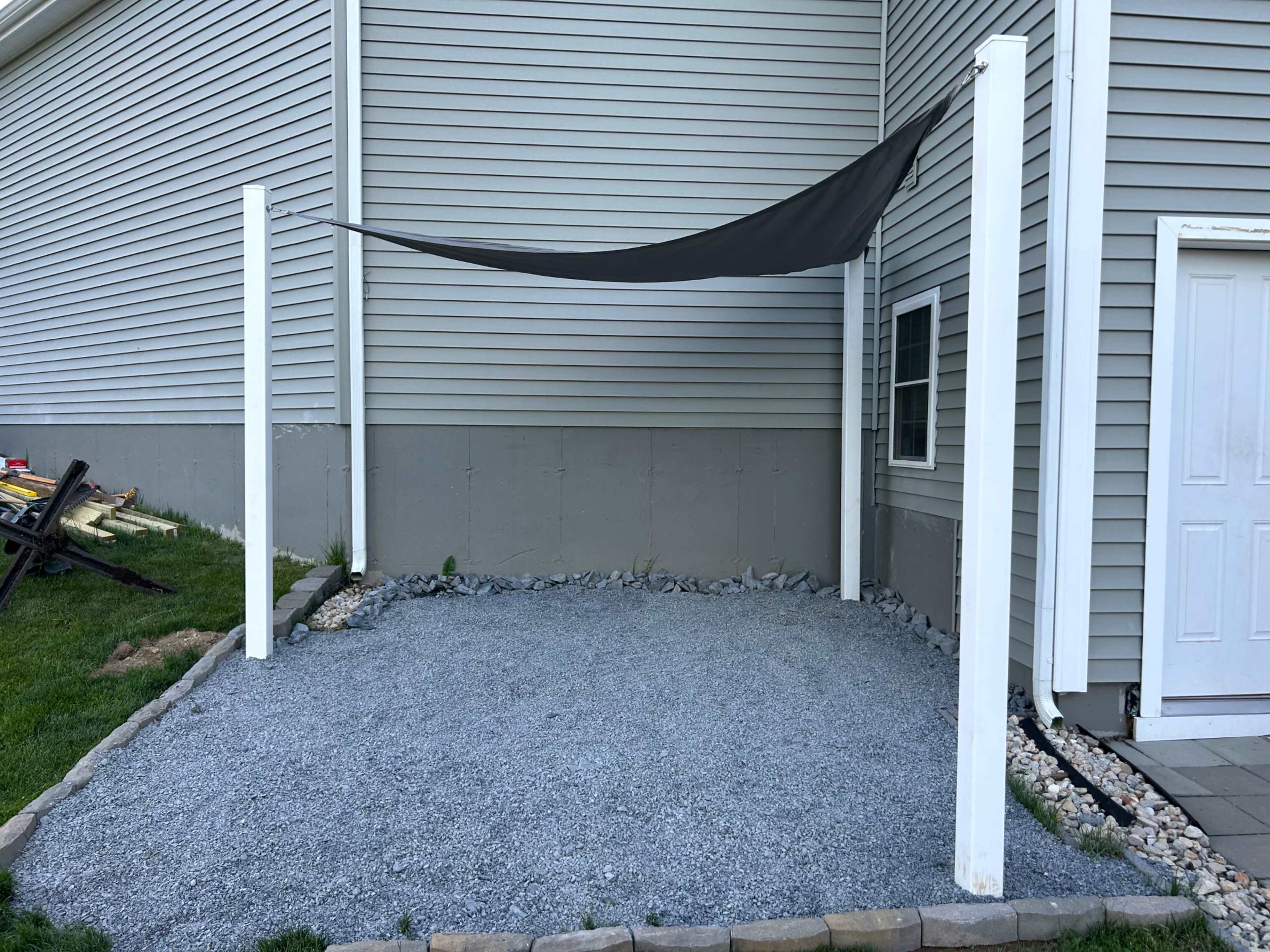 A gravel area with four white posts and a black shade sail, adjacent to a house with a gray exterior.