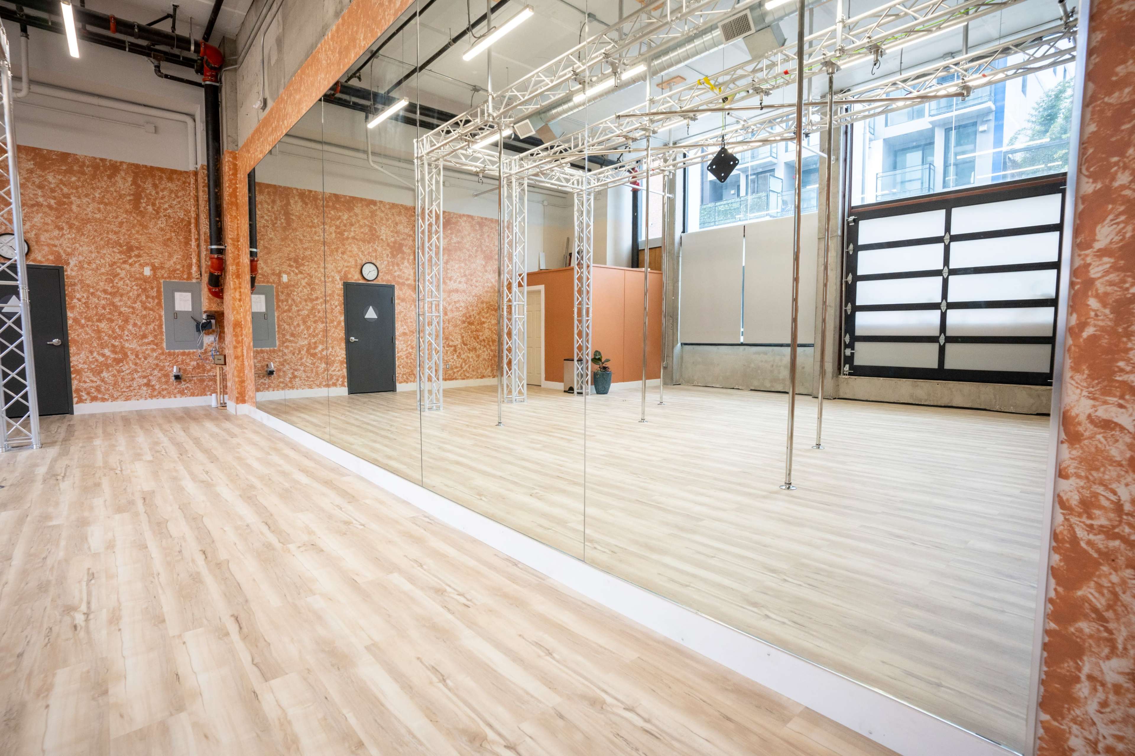 A spacious dance studio with wooden floors, mirrored walls, and a grid of metal trusses for lighting and equipment.