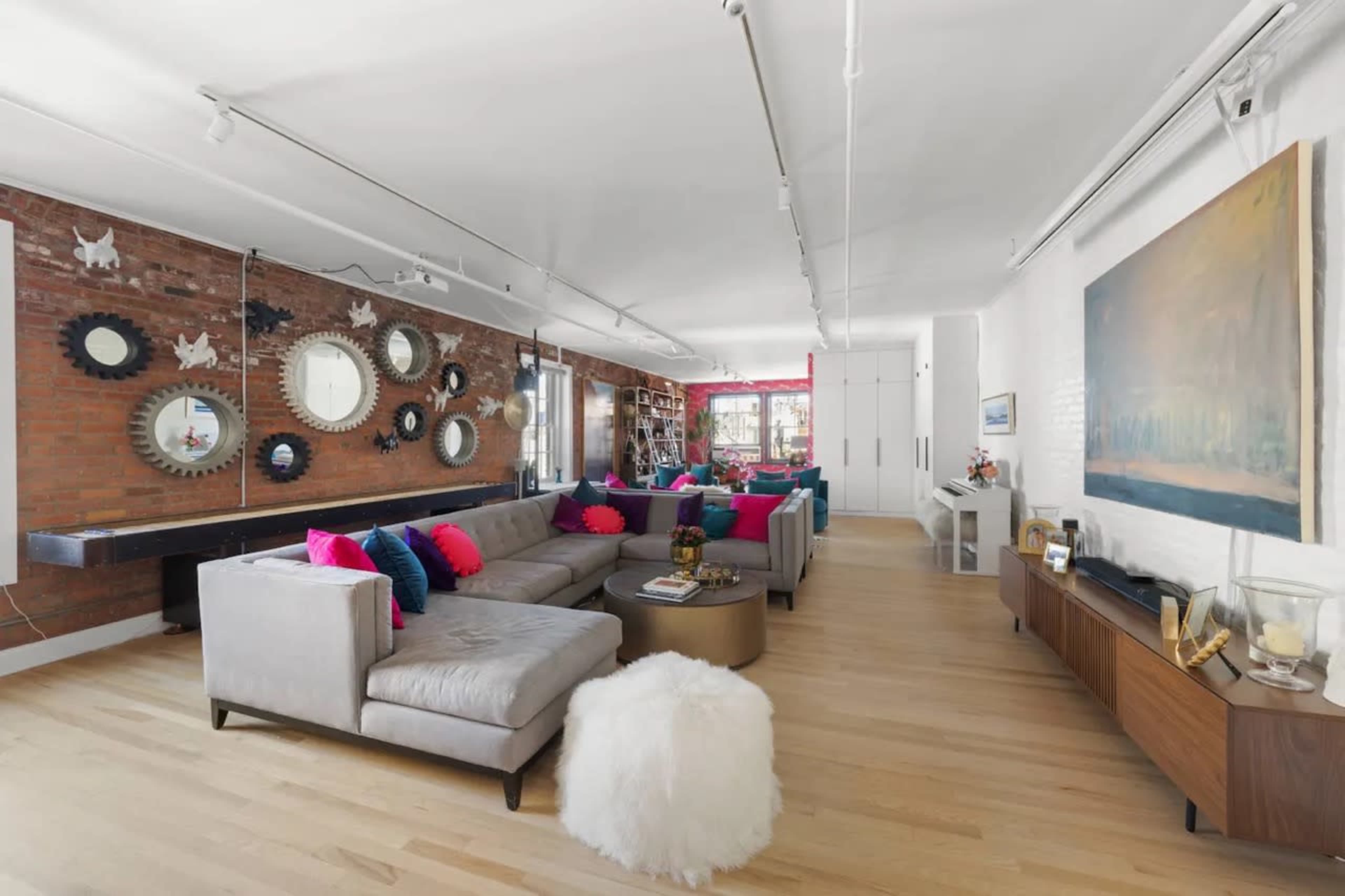 The image shows a spacious living room with a mix of modern furniture, colorful cushions, and a brick wall, featuring multiple mirrors and a large piece of artwork.