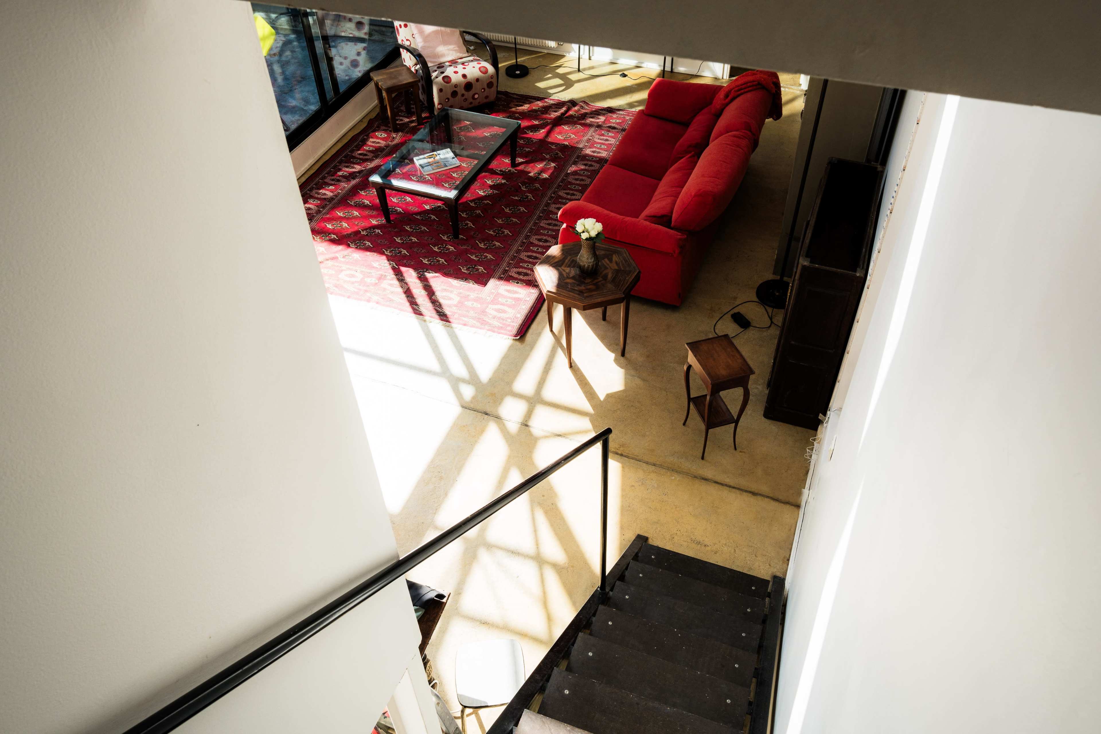 The image shows a modern living space viewed from an elevated angle, featuring a red sofa, a coffee table, and a patterned rug, with stairs leading down to the area.