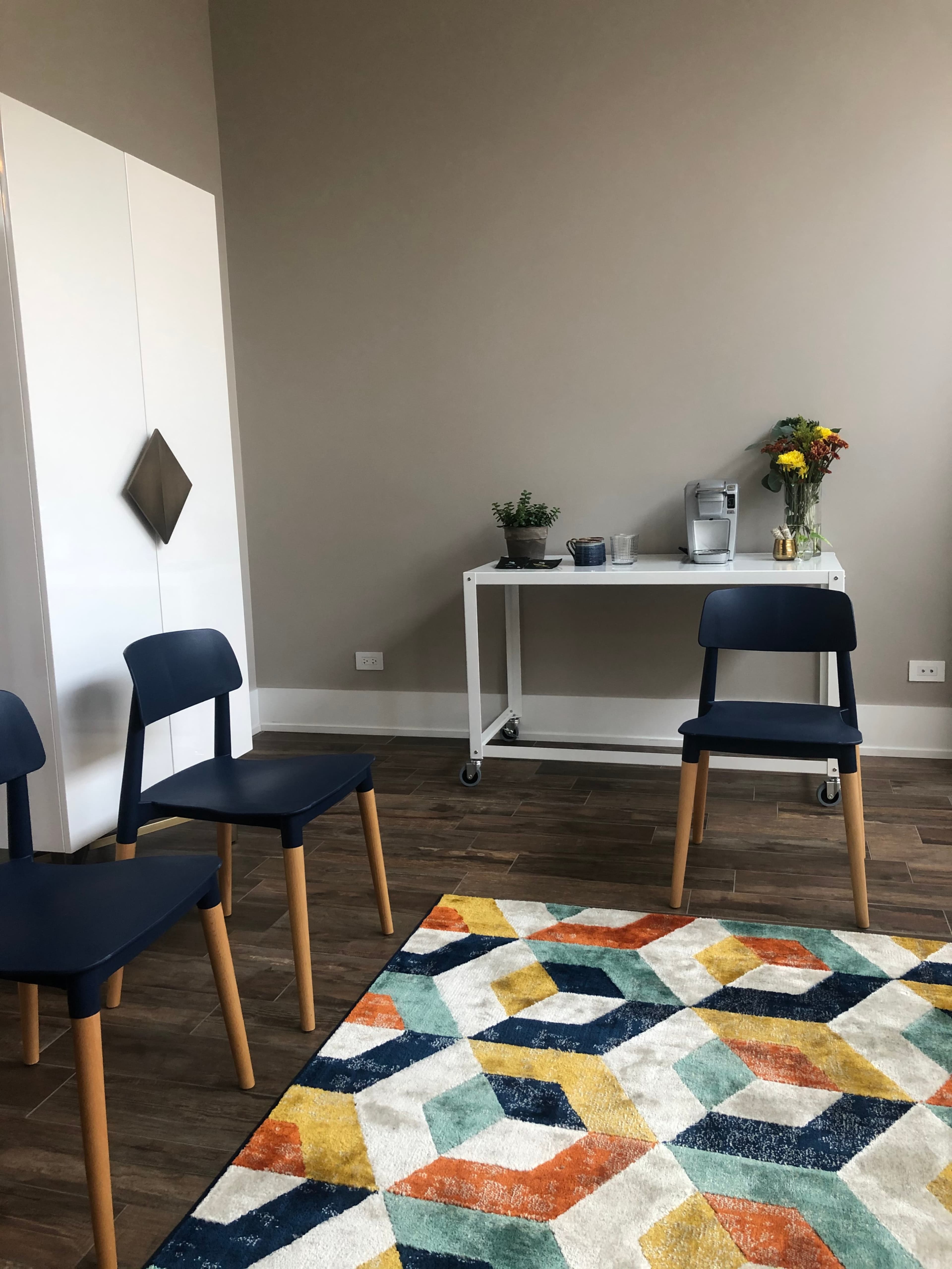 A minimalist room with three blue chairs arranged around a geometric rug, a white cart with a coffee maker and flowers, and a blank wall.