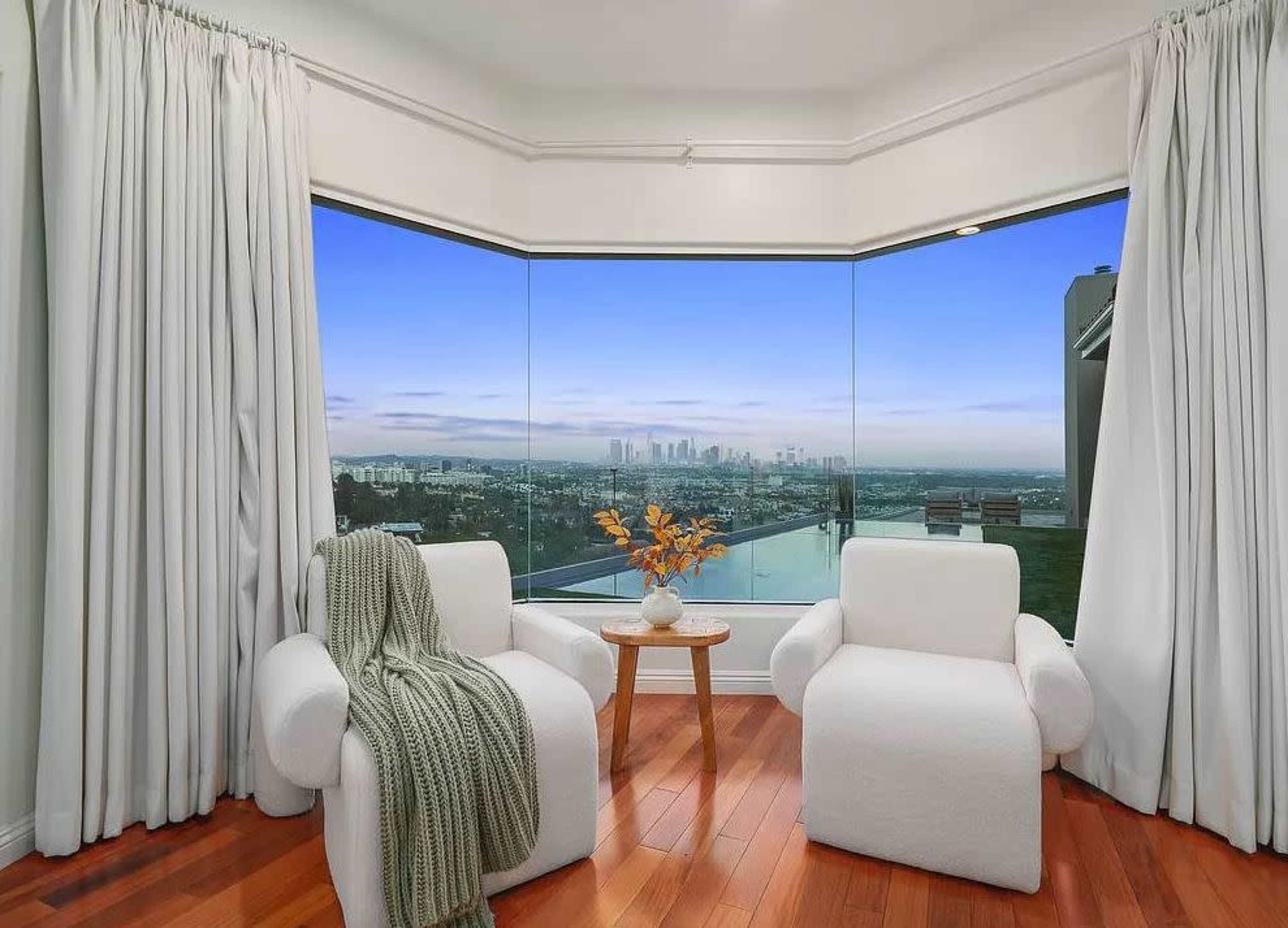 A cozy living area features two white armchairs and a small wooden table with a vase of dried flowers, framed by large windows showcasing a view of the city skyline.