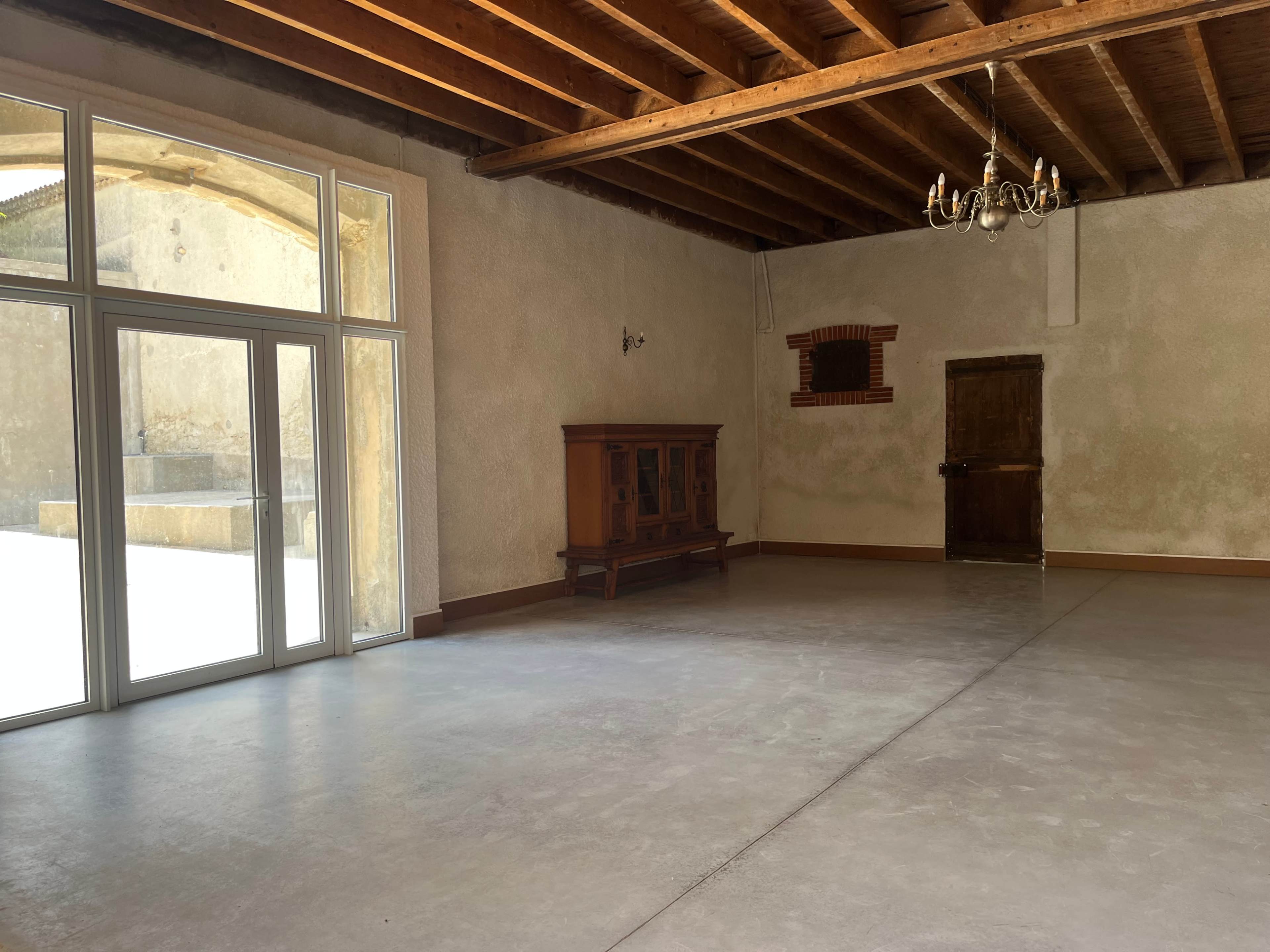The image shows a spacious, empty room with a wooden ceiling, large windows, a chandelier, and a wooden cabinet against the wall.