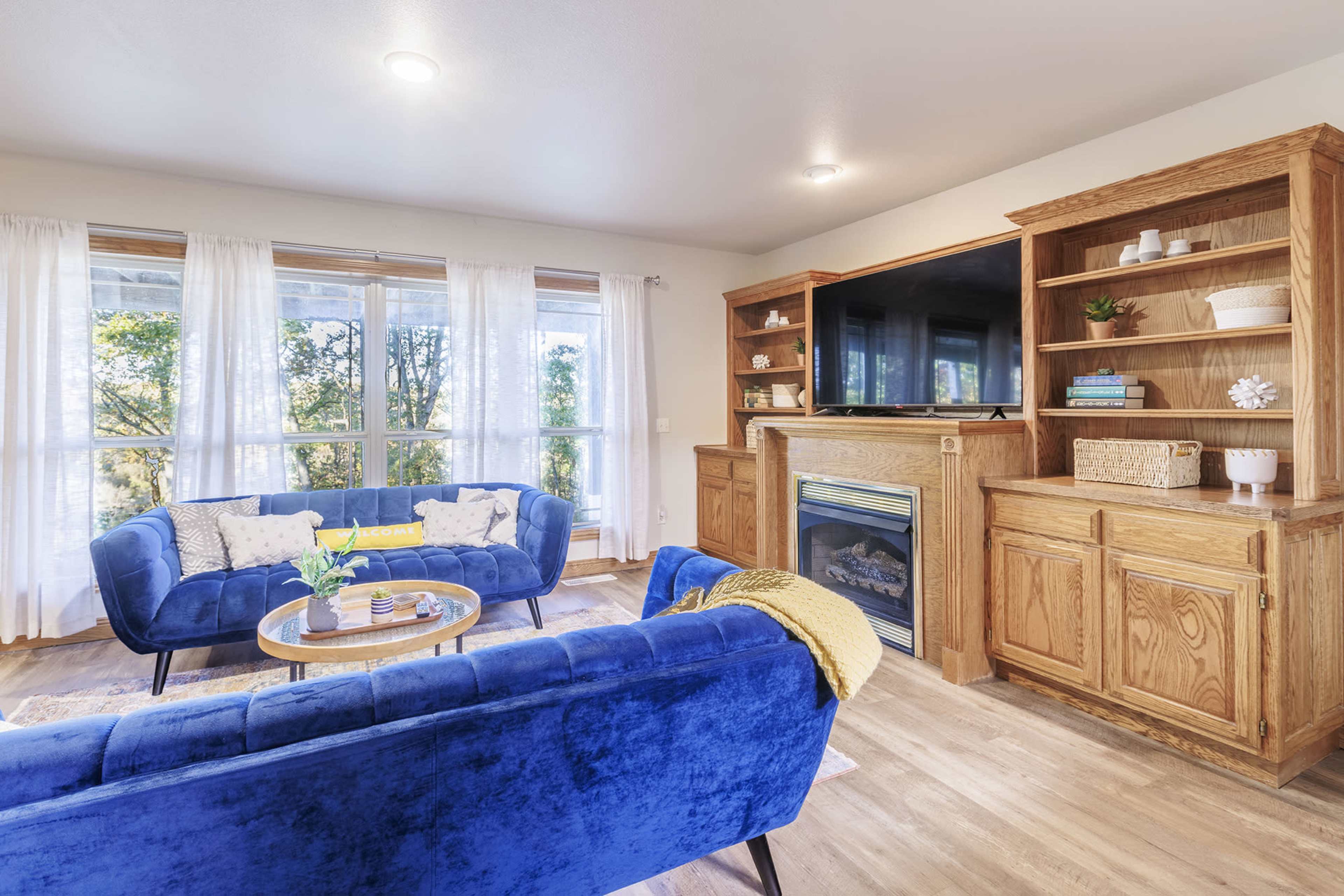 A cozy living room features two blue sofas, a wooden entertainment center with a TV, and large windows with light curtains that overlook a green view.