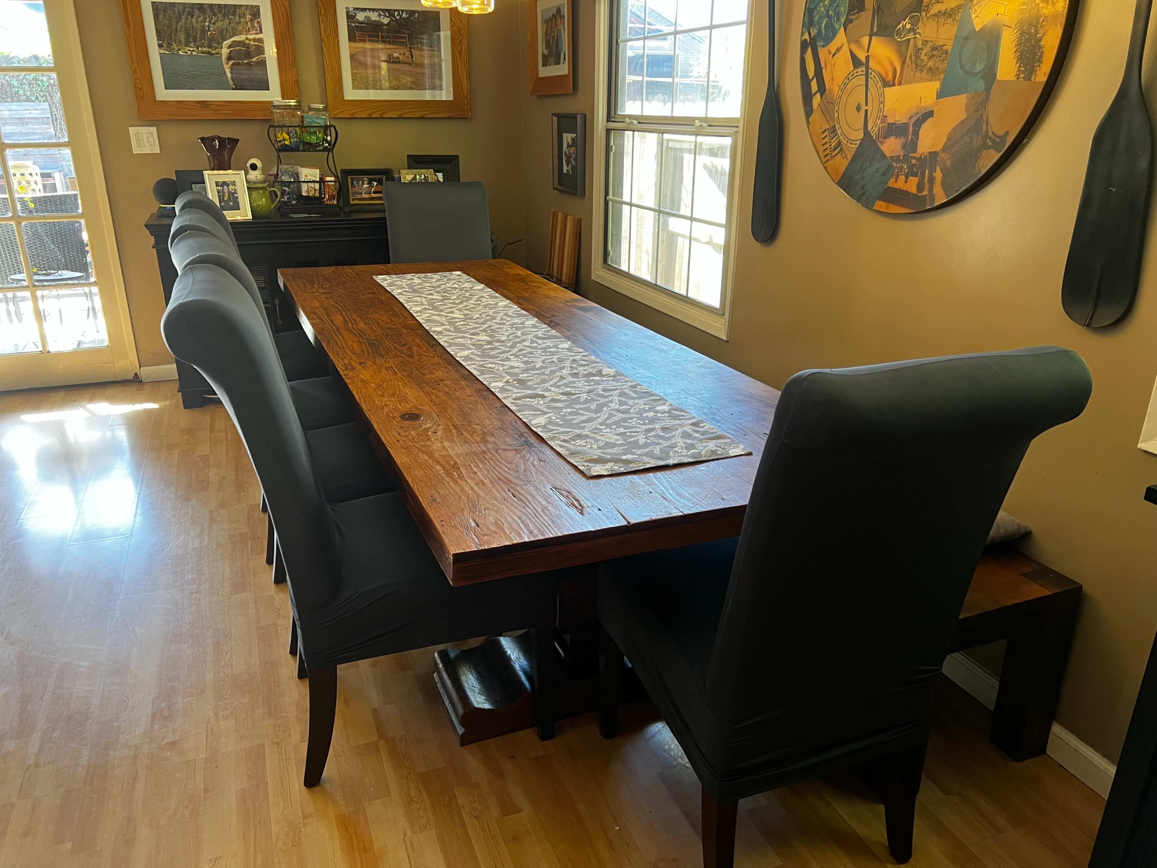 A long wooden dining table with a patterned runner is set in a well-lit room featuring several framed photographs and a large circular wall decoration.