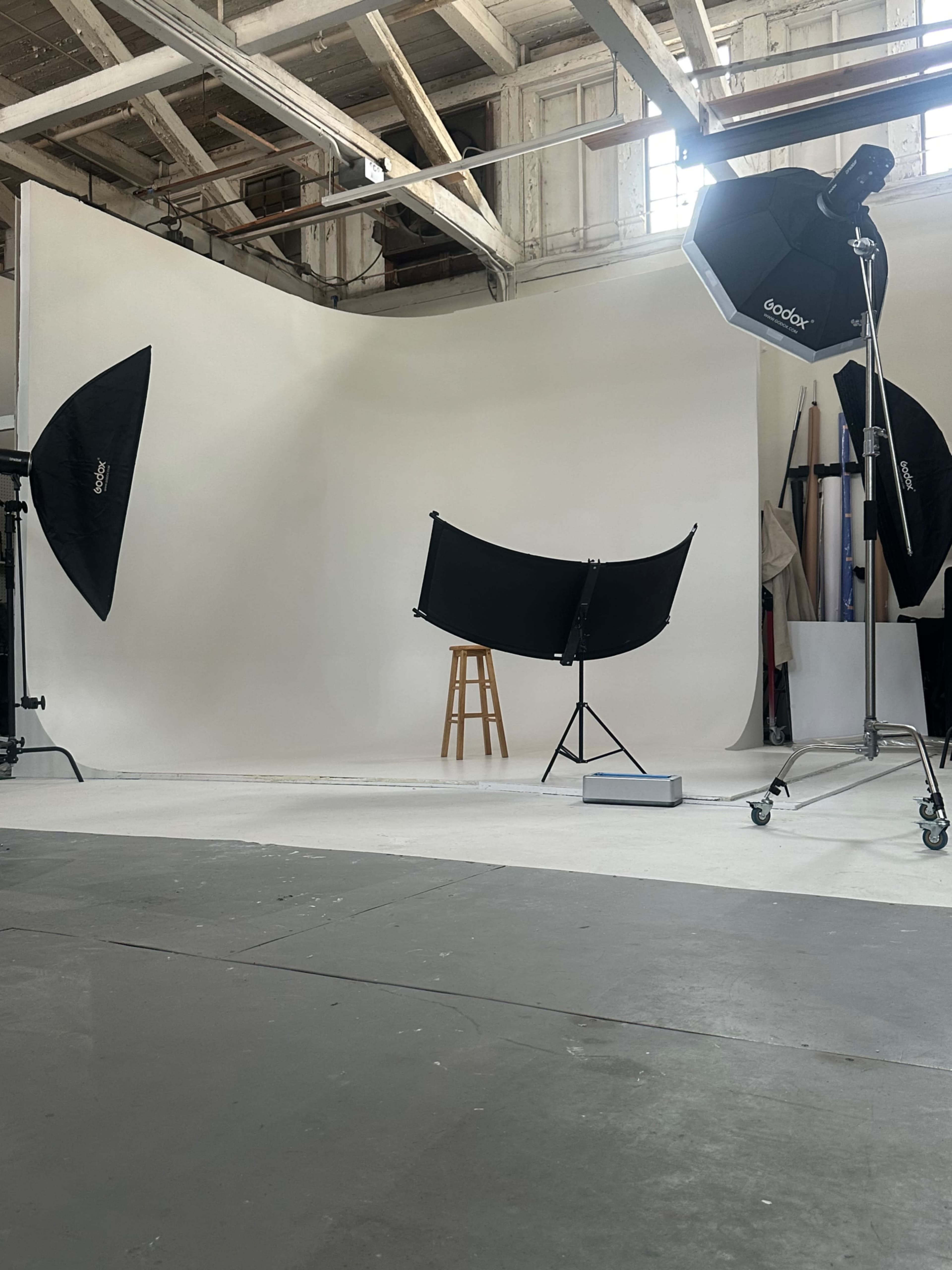 The image shows a photography studio with a plain backdrop, a wooden stool, and studio lighting equipment positioned around the setup.