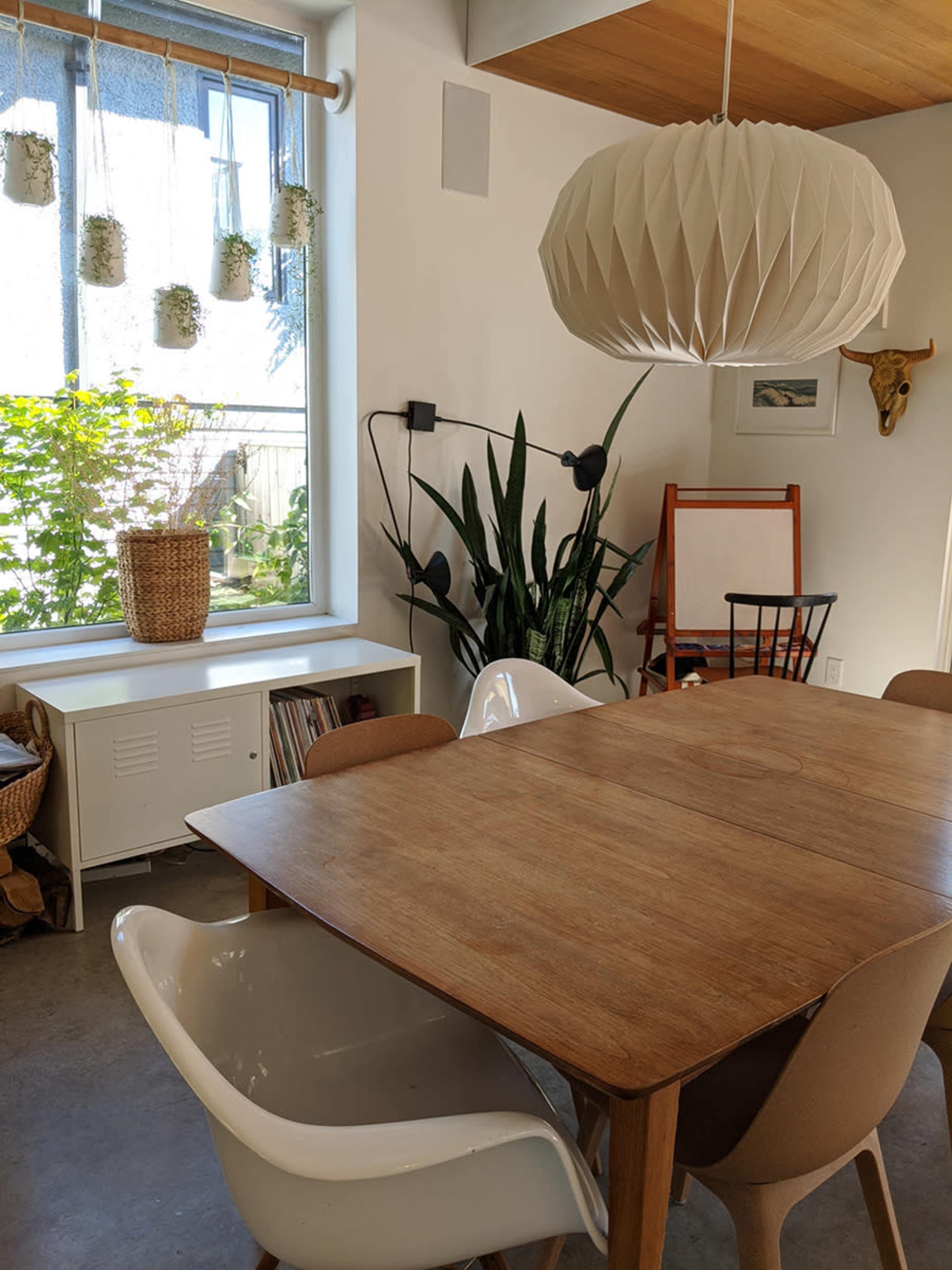 A modern dining area features a wooden table surrounded by various chairs, with a large pendant light overhead and a window showcasing greenery outside.