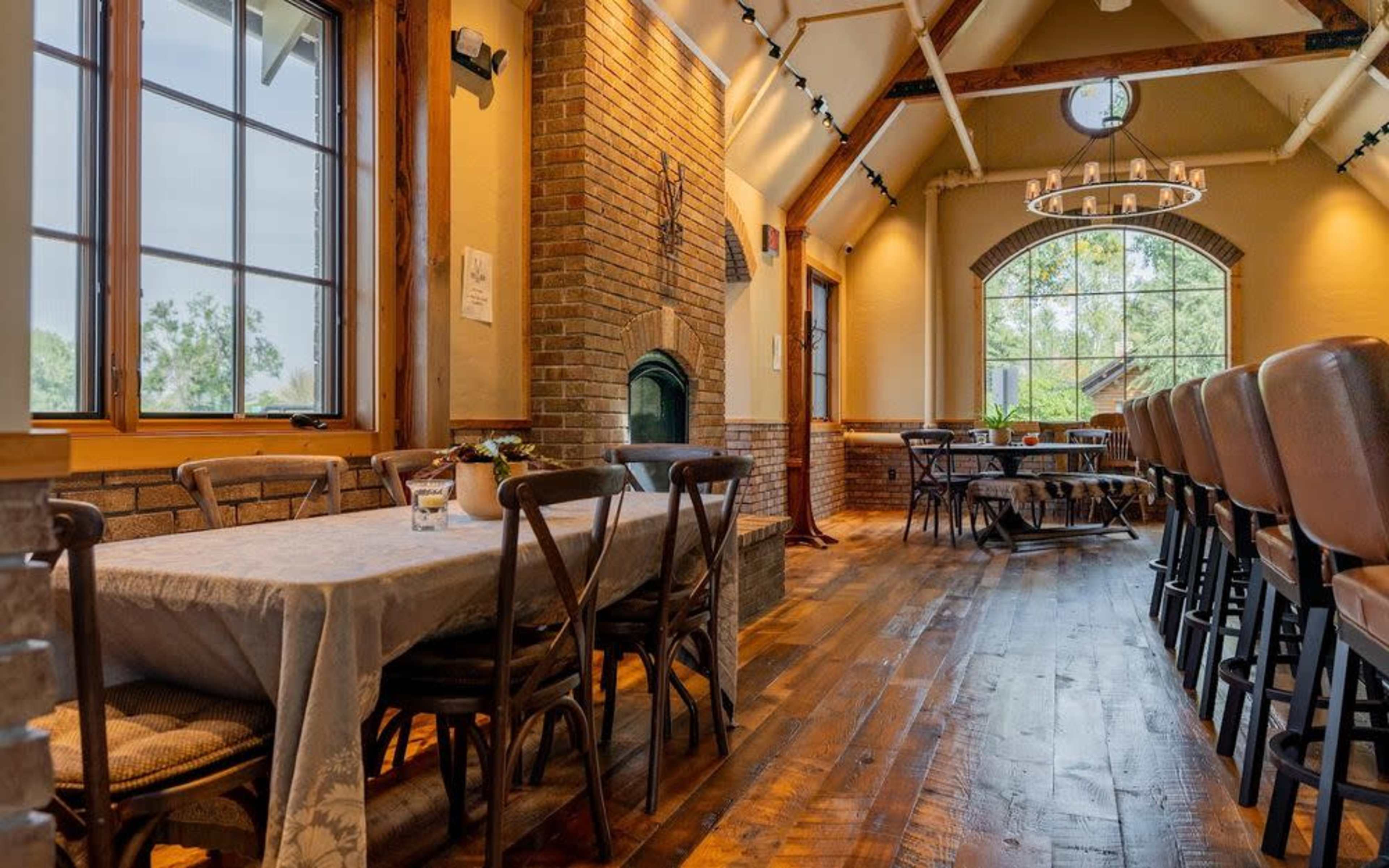 The image shows a spacious dining area with wooden tables and chairs, brick walls, and a large window allowing natural light to enter.