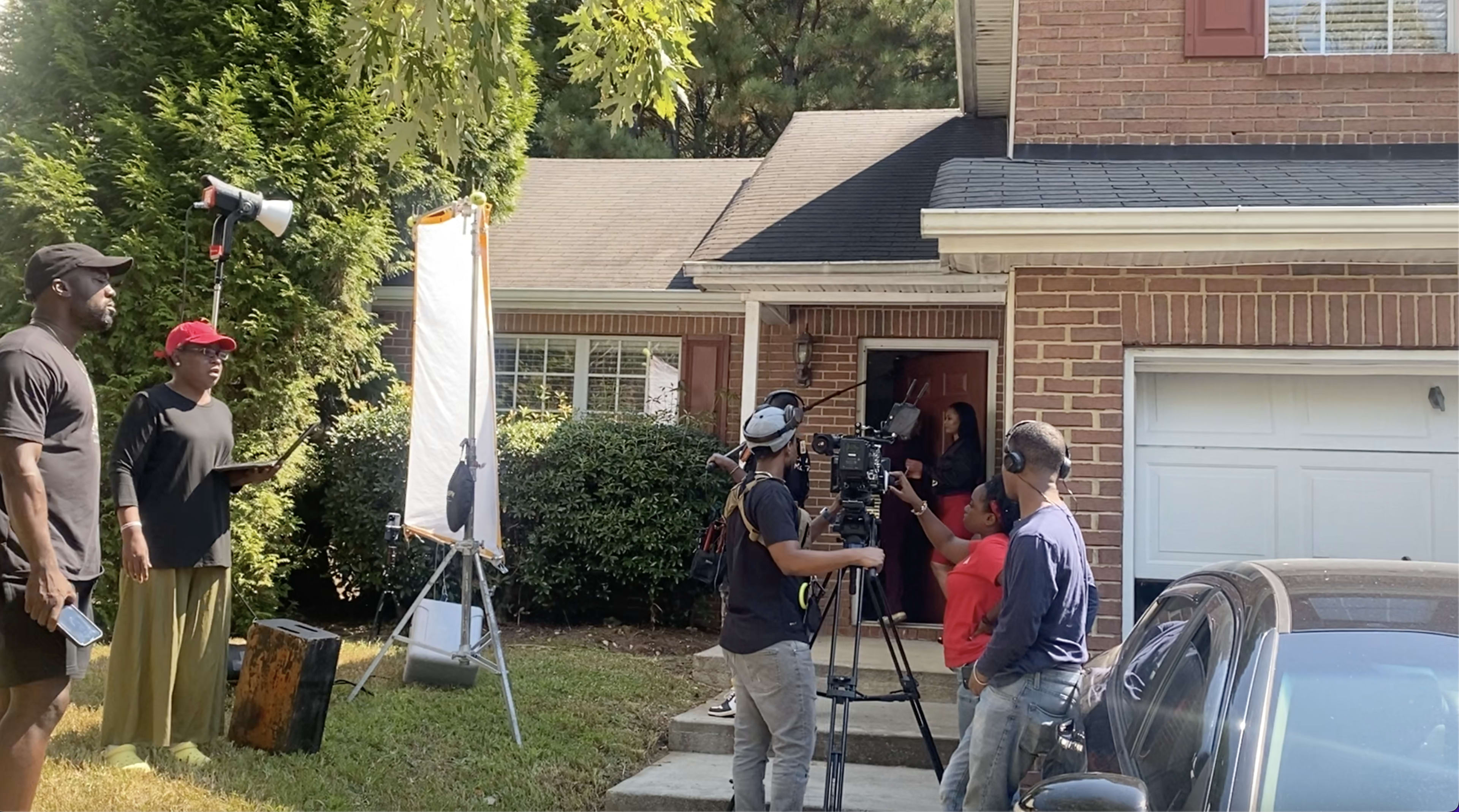A film crew sets up equipment outside a brick house as a woman stands in the doorway.