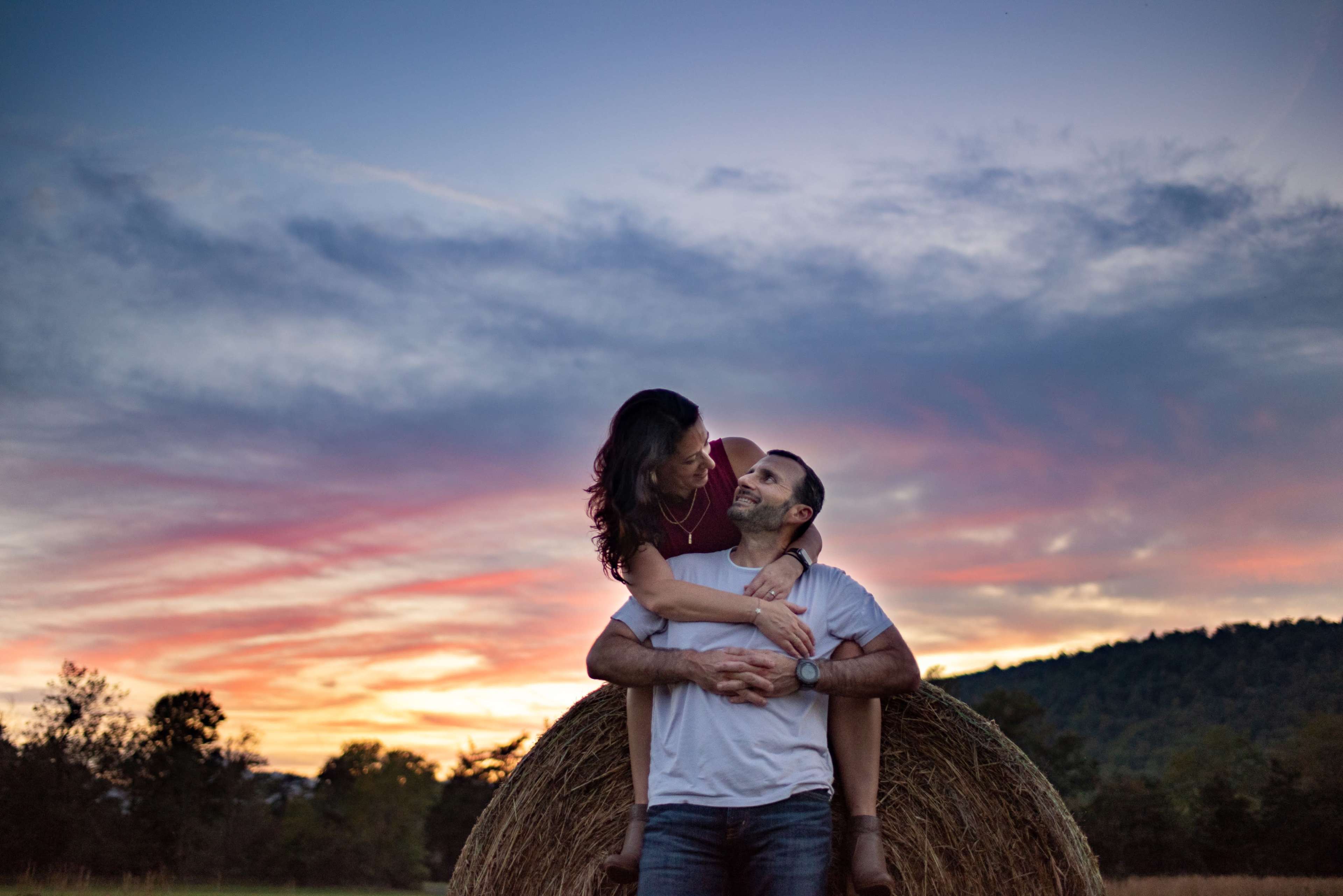 A couple poses on a hay bale against a sunset sky with colorful clouds and a forested hill in the background.