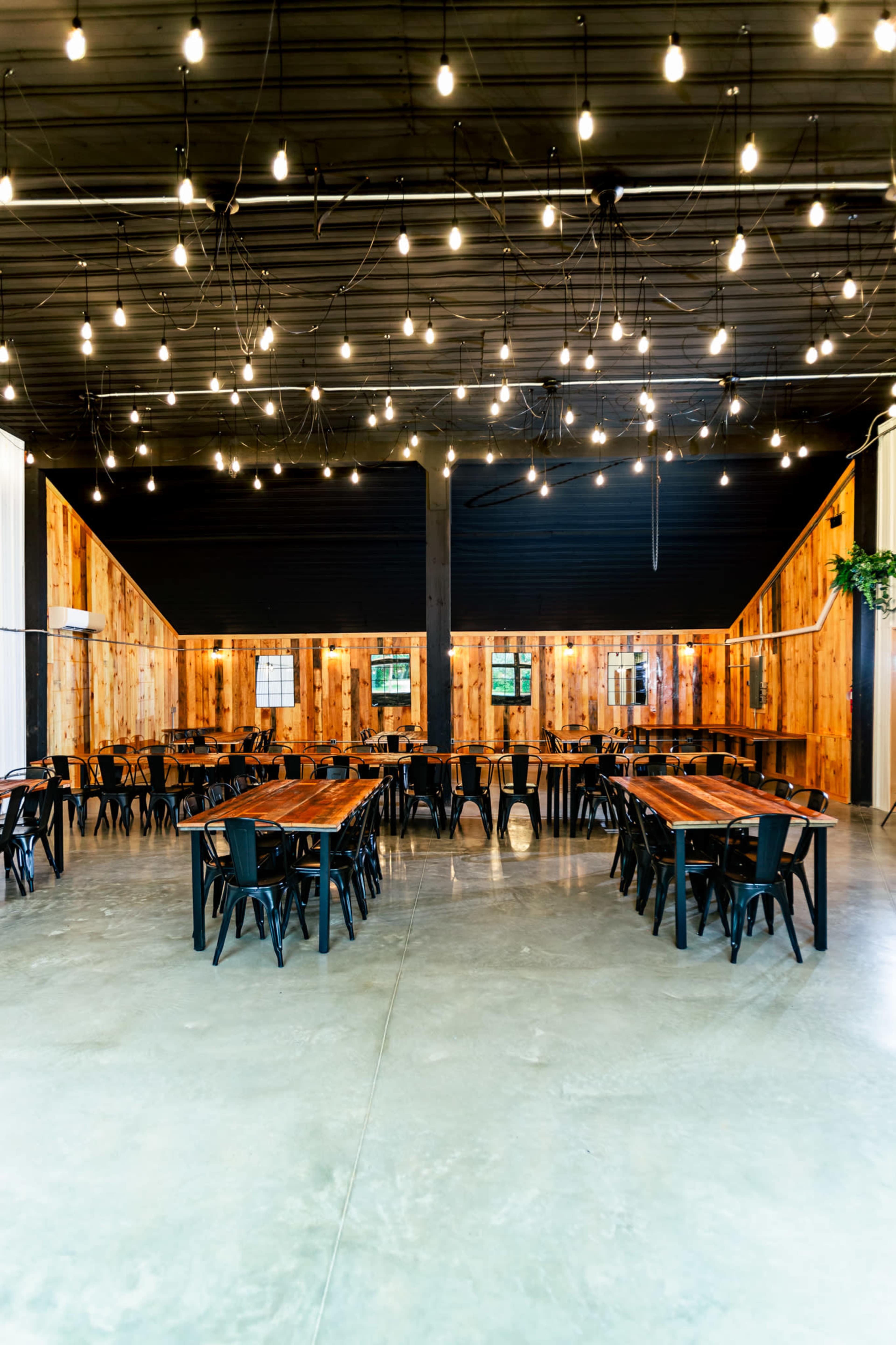 The image shows a spacious dining area with multiple wooden tables arranged in a symmetrical layout, surrounded by wooden walls and illuminated by hanging string lights.