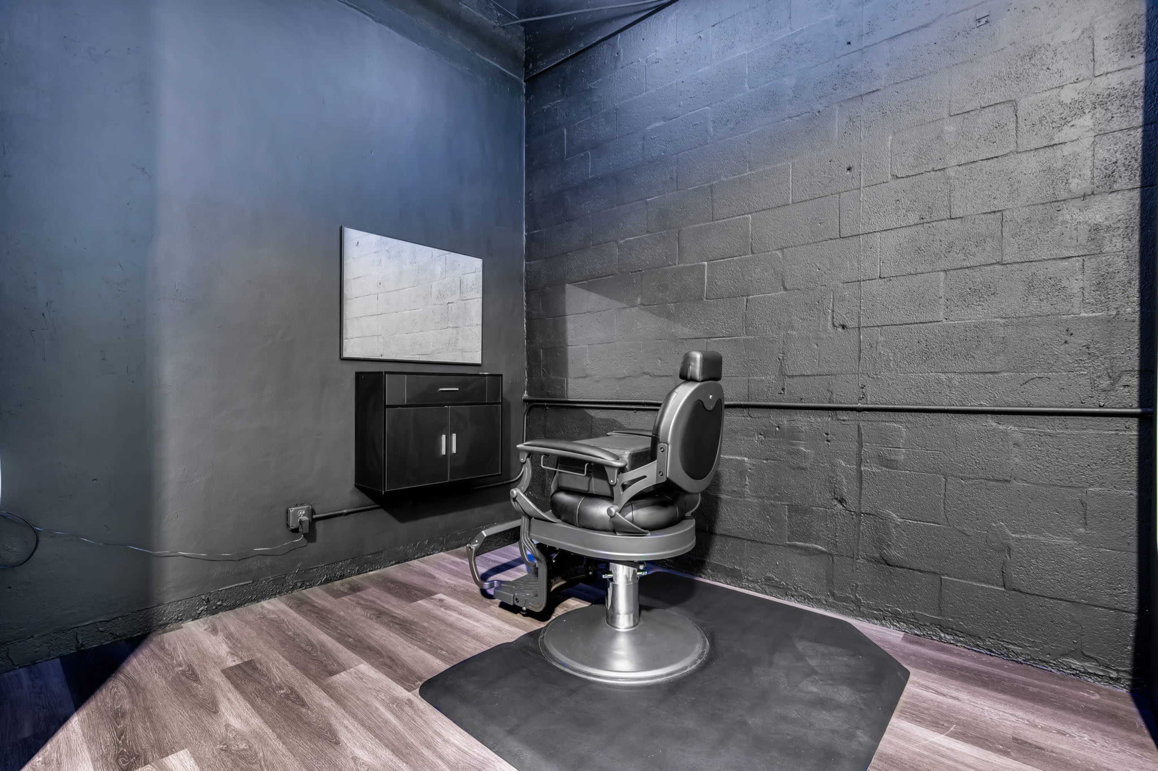 The image shows a minimalistic barber or salon space with a black wall, a black styling chair on a gray mat, and a small mirror mounted on the wall.