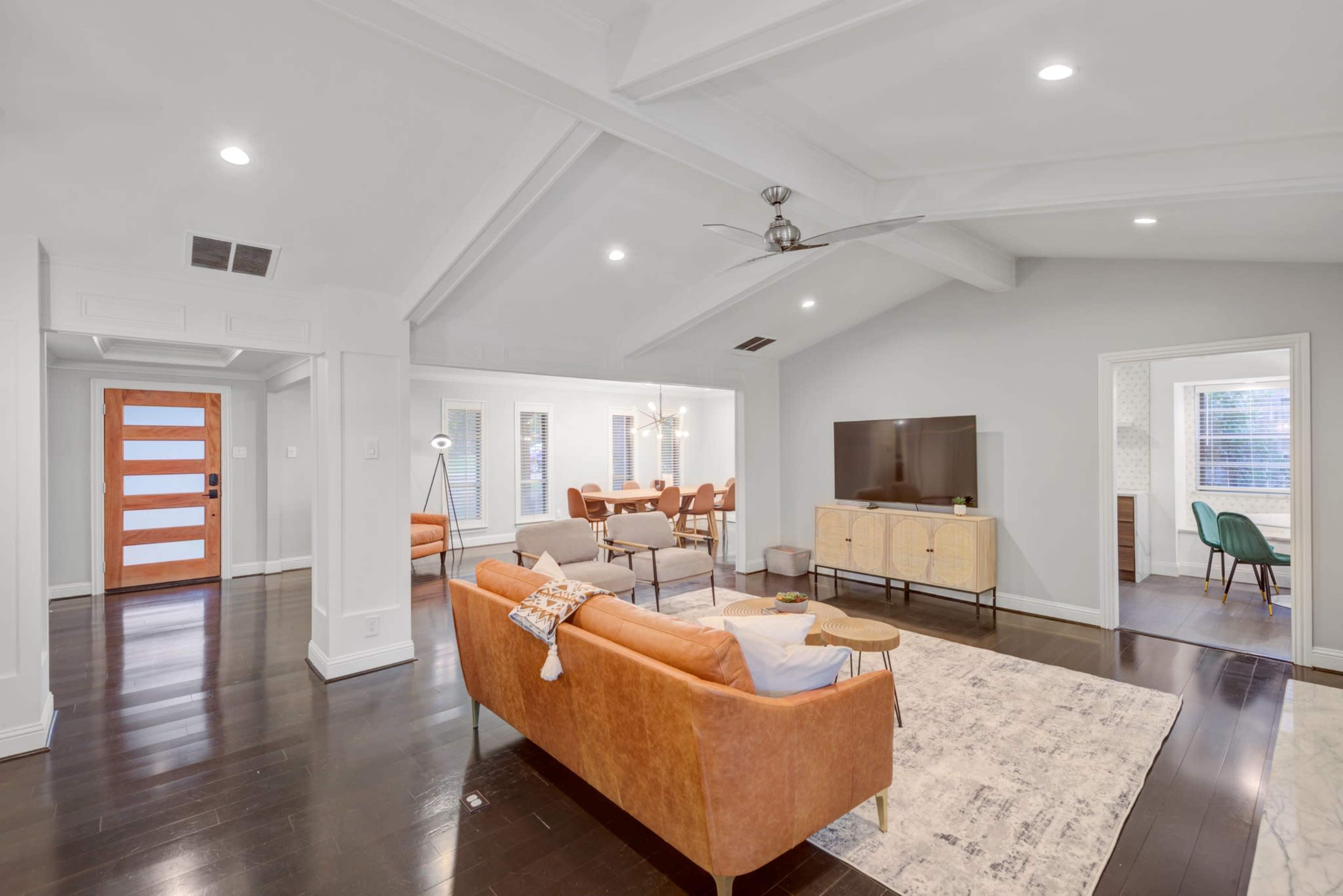 The image shows a modern, open-concept living room with a sofa, coffee table, and television, connected to a dining area and featuring large windows and wooden floors.