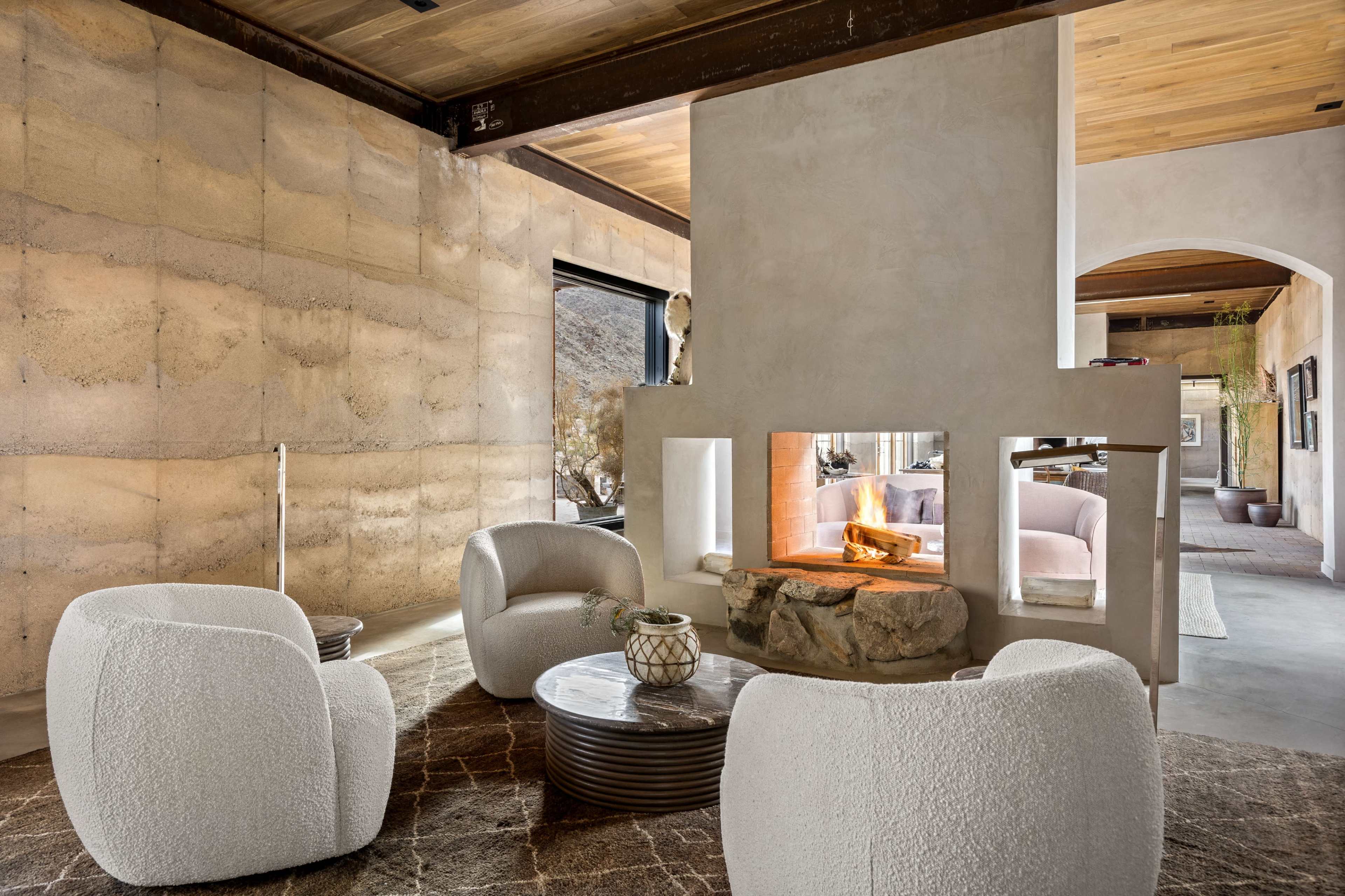 The image shows a modern living room with a central stone fireplace, featuring four rounded white chairs arranged around a circular table on a textured rug.