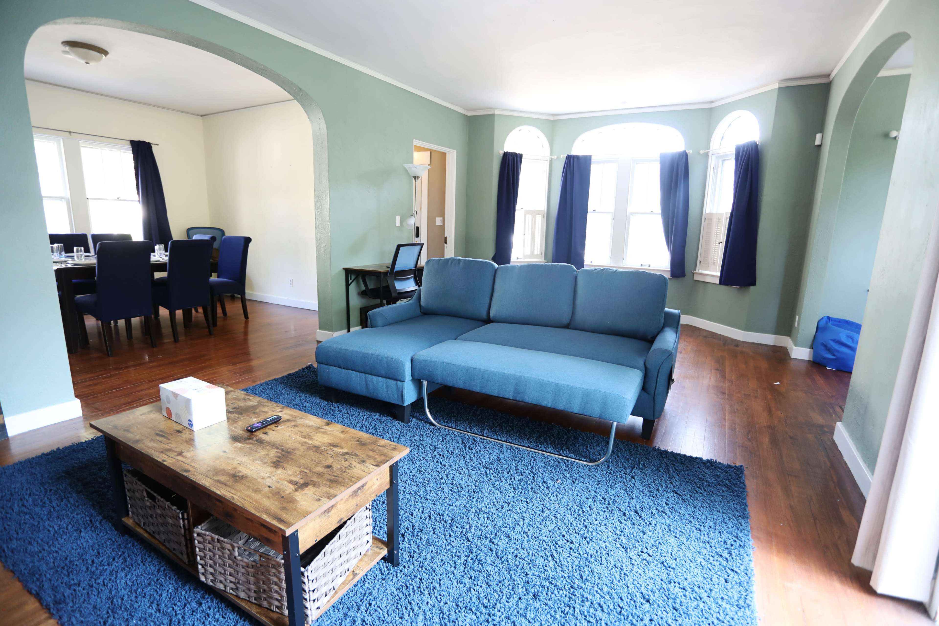 The room features a blue sectional sofa, a wooden coffee table, and a textured area rug, with windows allowing natural light to enter.