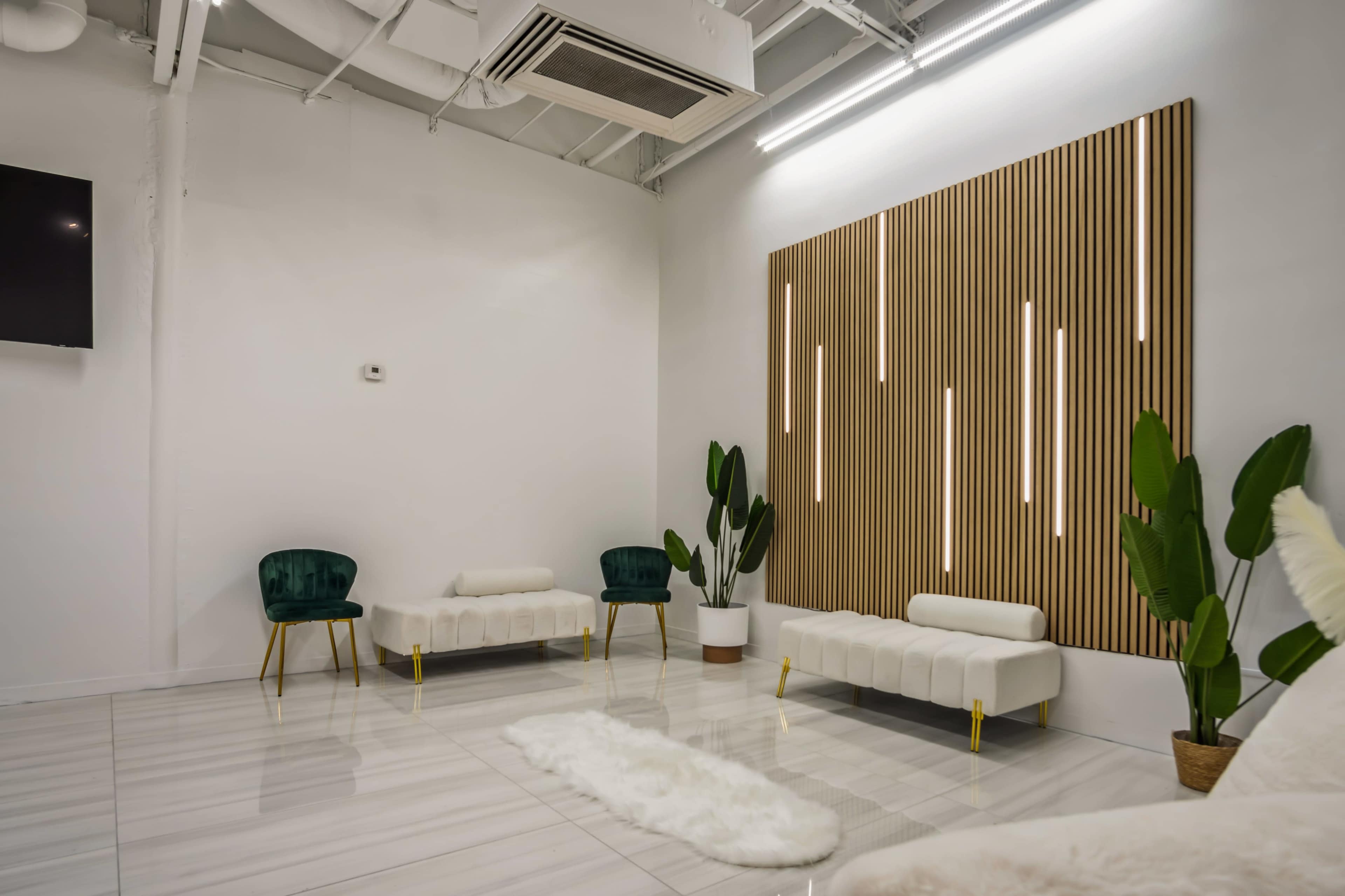 The image shows a modern, minimalist interior space with green chairs, a white rug, and a wood-paneled accent wall featuring vertical light strips.