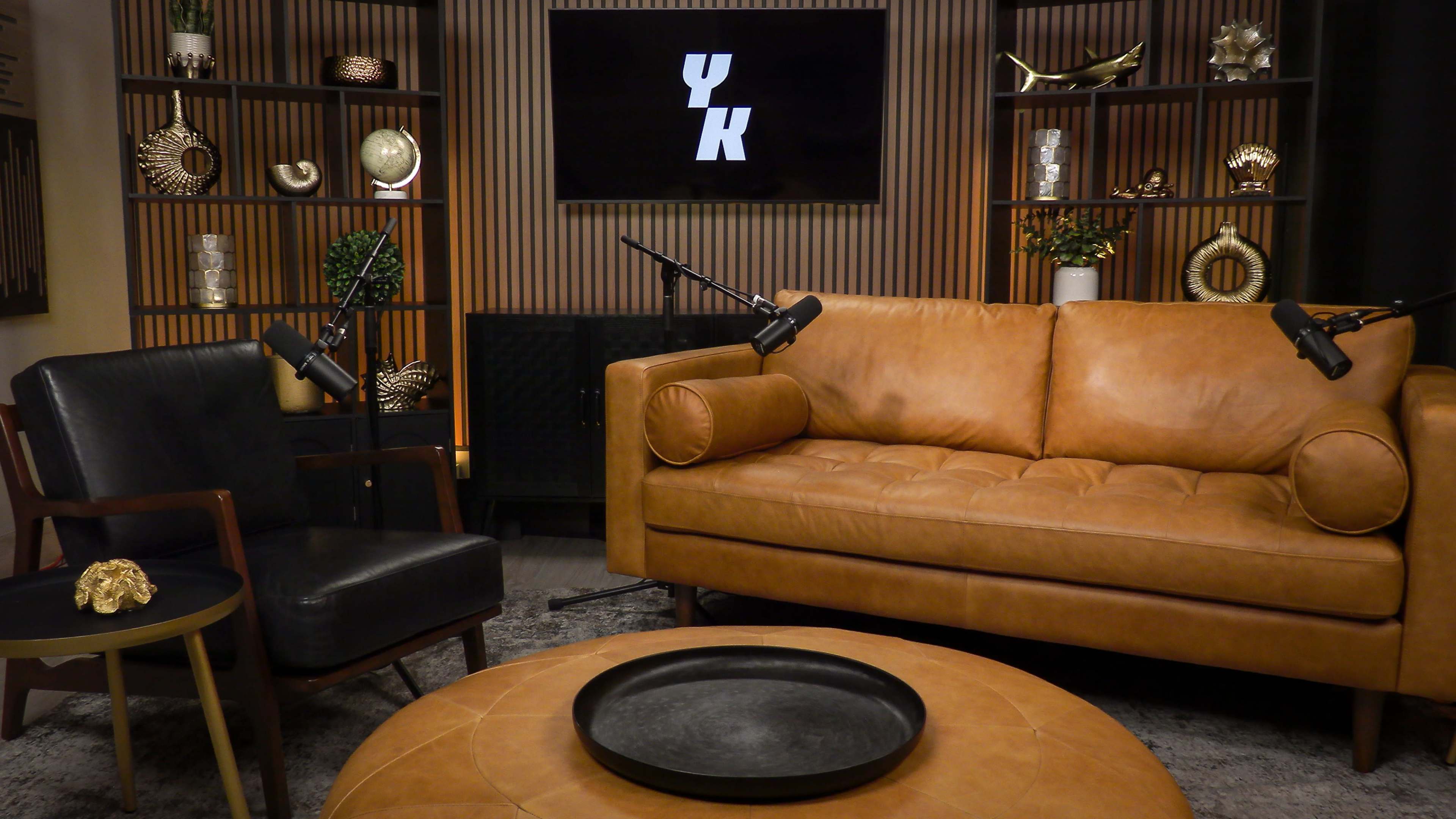 The image shows a modern chat or interview set featuring a brown leather sofa, a black armchair, a round coffee table, and a backdrop with a logo on a screen surrounded by decorative shelves.