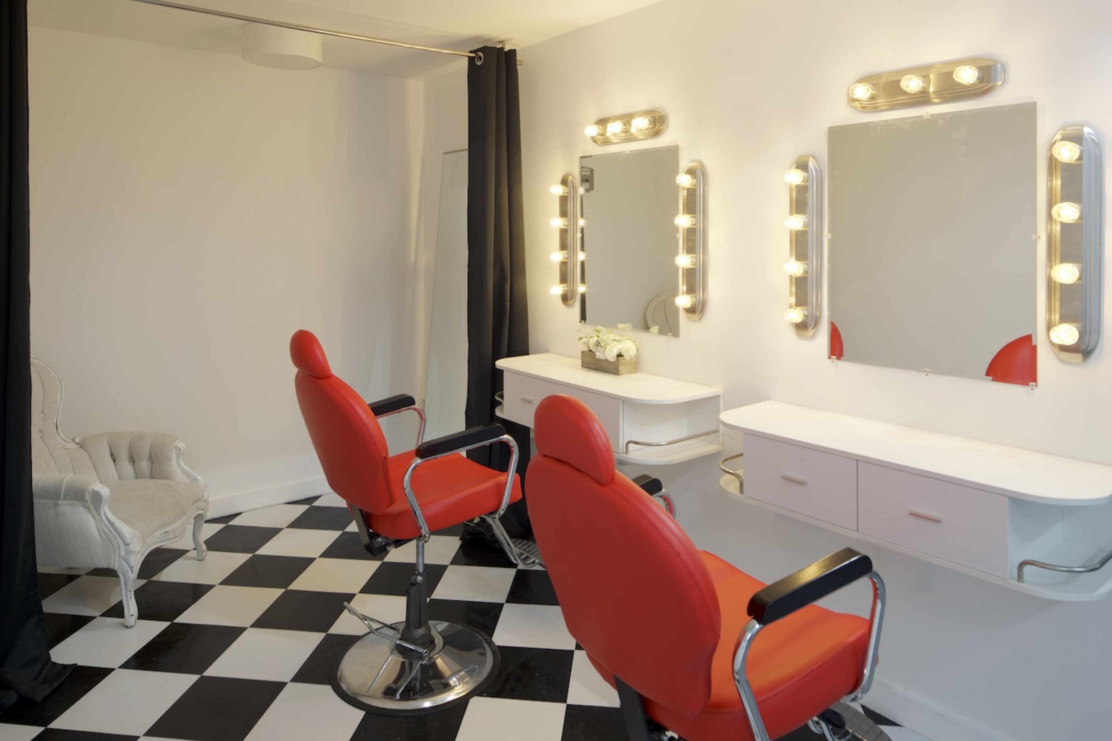 The image shows a hair salon with two red barber chairs, a checkered floor, and illuminated mirrors against a white wall.