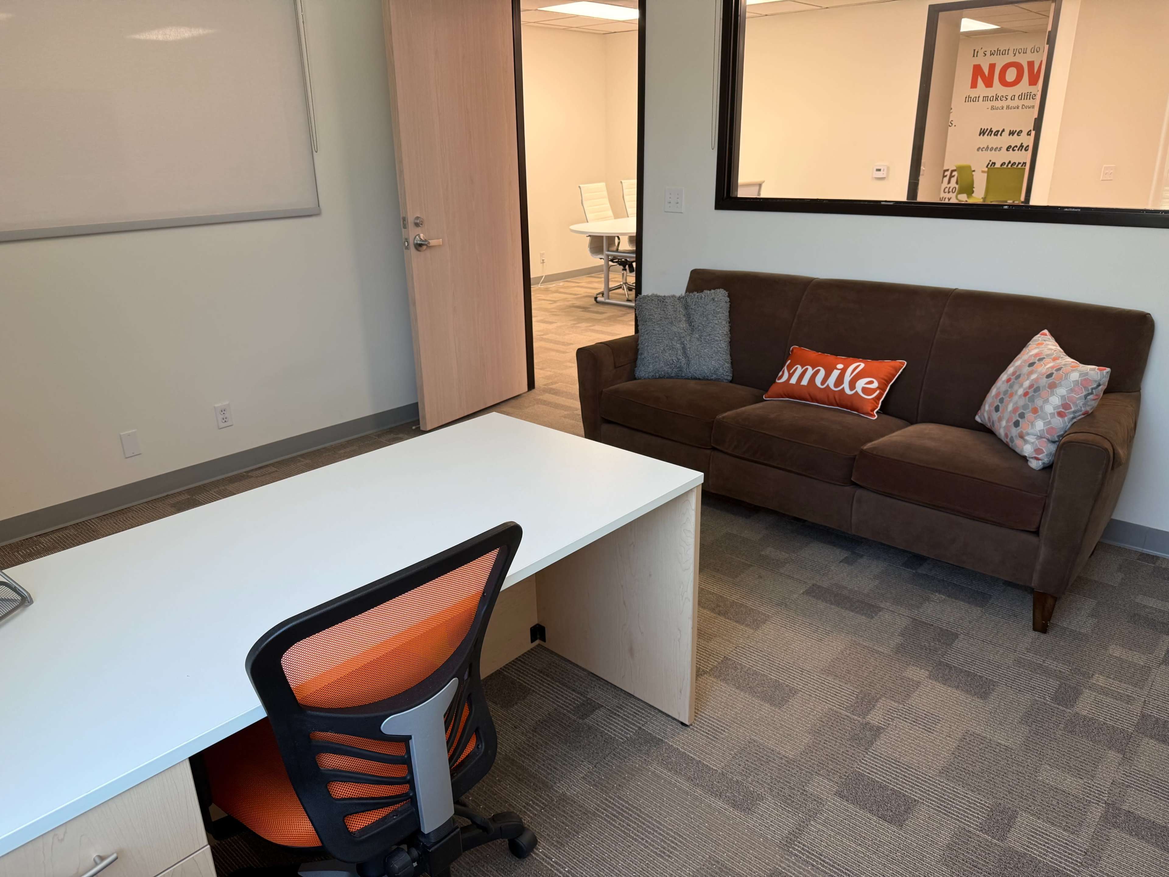 The image shows a small office space featuring a desk with an orange and black office chair, a brown sofa with decorative pillows, and a window overlooking another meeting area.