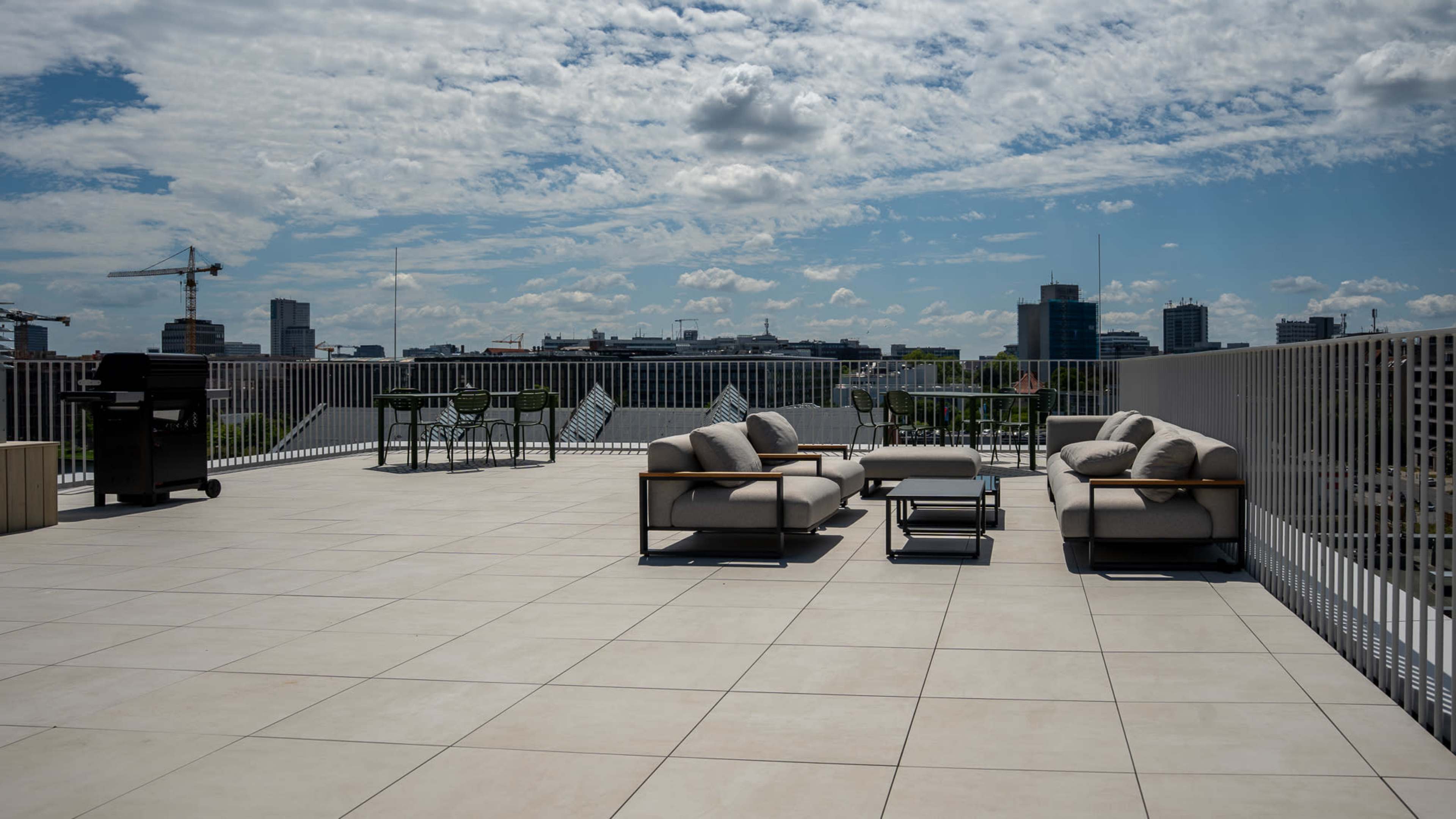 The image shows a spacious rooftop terrace featuring modern outdoor furniture, including sofas, a coffee table, and a dining set, with a cityscape in the background.