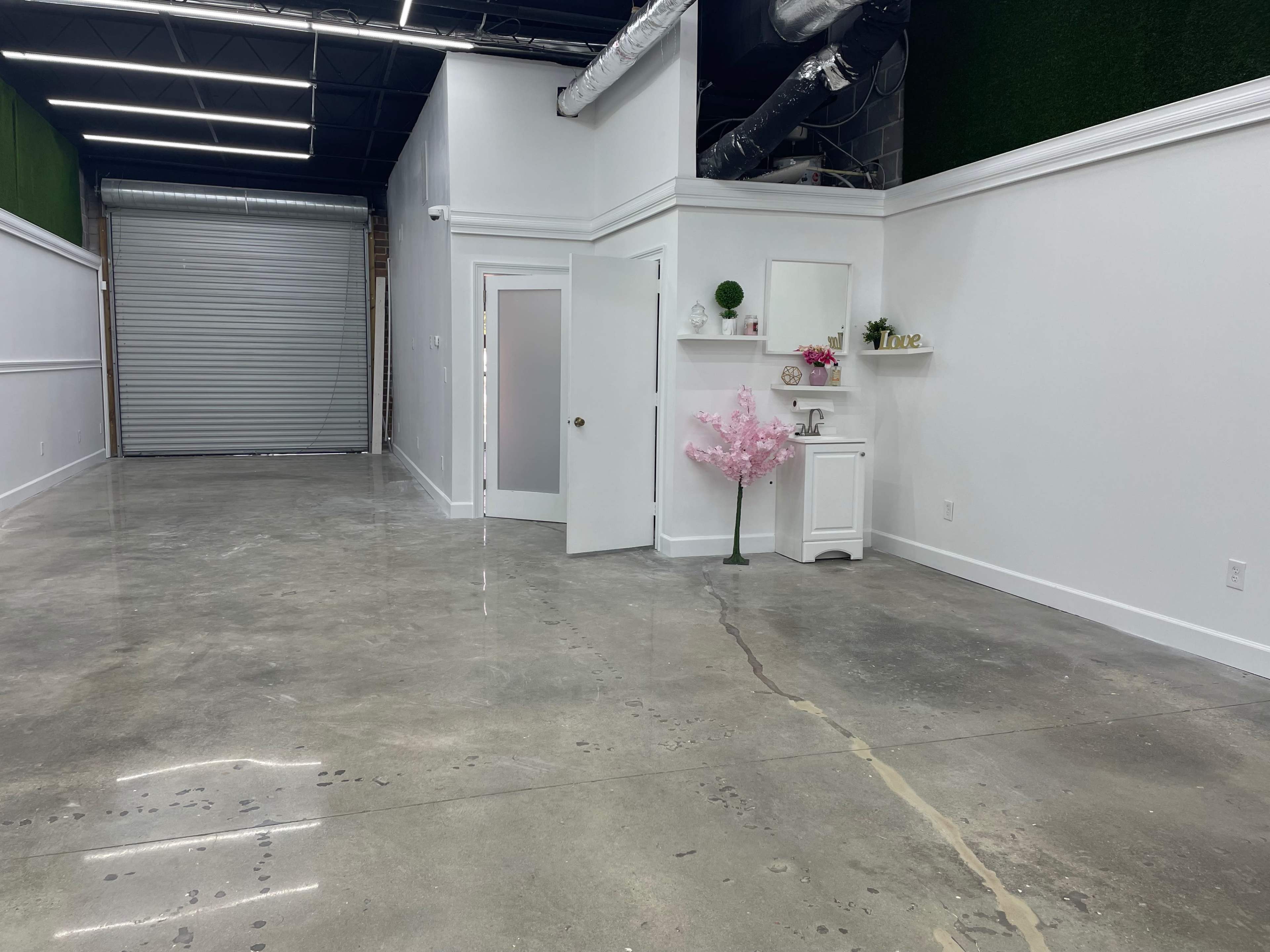An empty, well-lit retail space with polished concrete floors, a door leading to another room, and a small white cabinet decorated with artificial flowers against the wall.