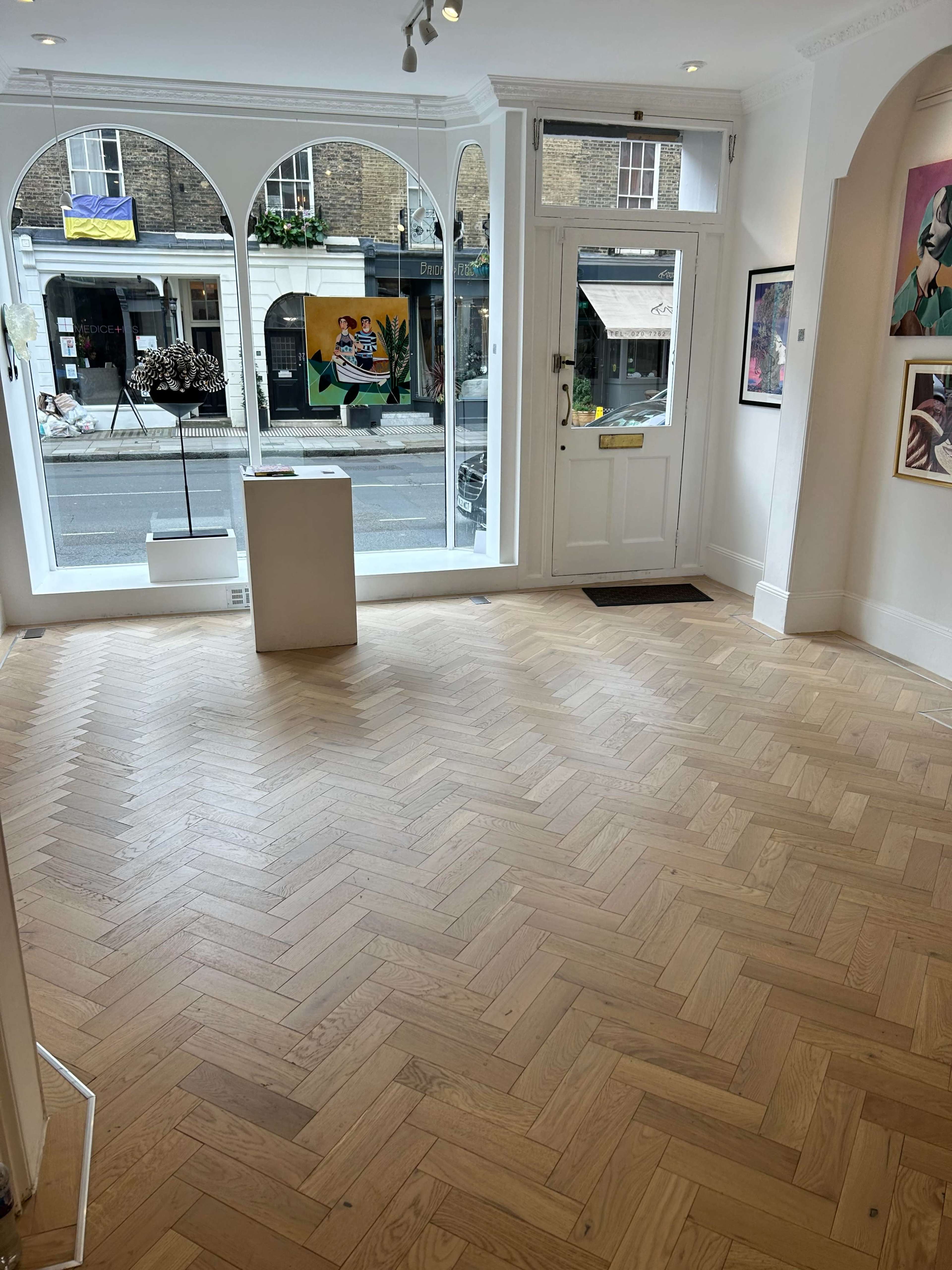 A gallery interior with herringbone-patterned wooden flooring and large windows displaying a street view outside.