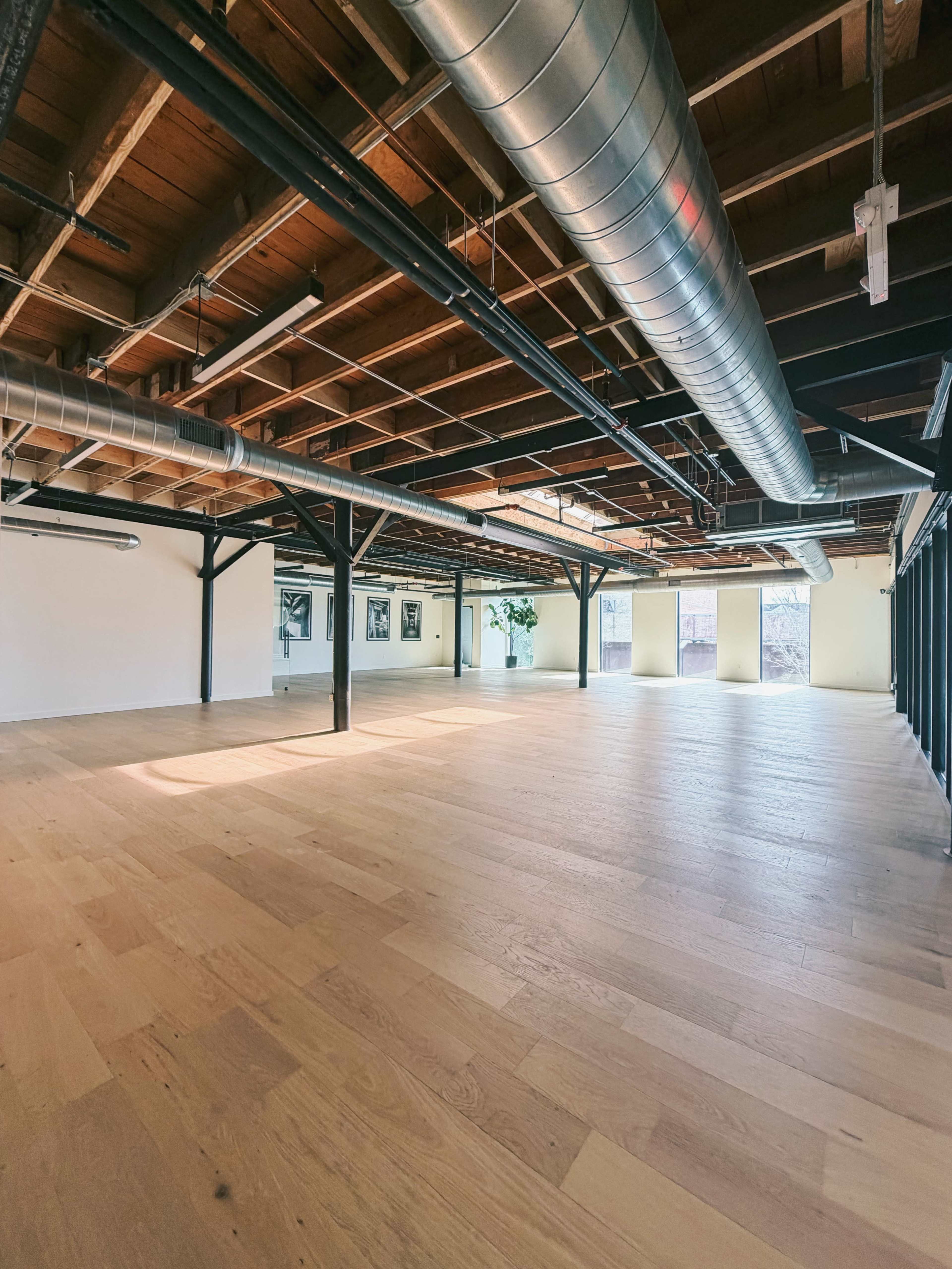 The image shows an empty, spacious interior of a commercial building with wooden flooring, exposed beams, and ductwork overhead.