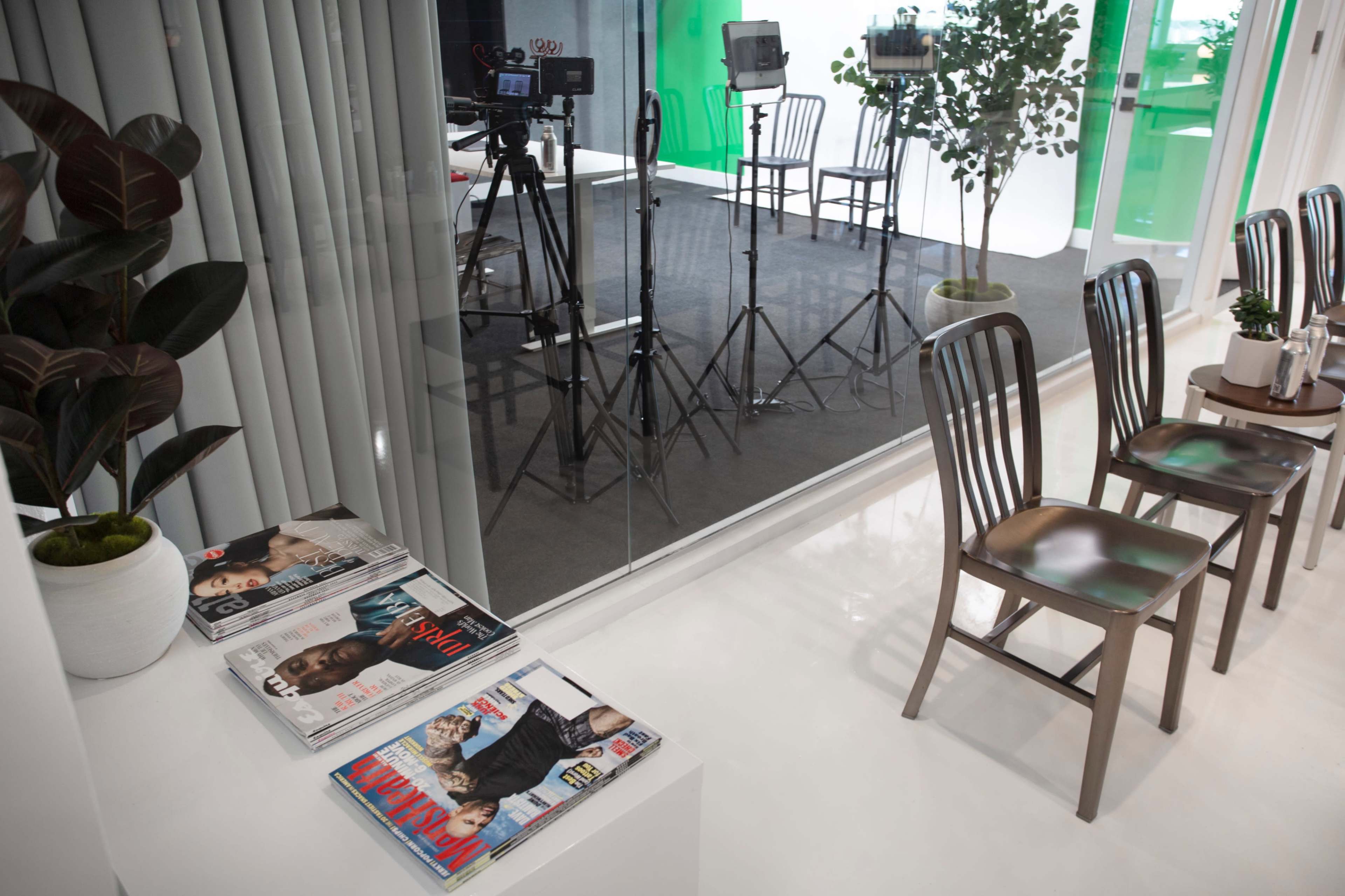 A minimalist studio space features rows of empty chairs, a shelf of magazines, and cameras set up against a glass wall.