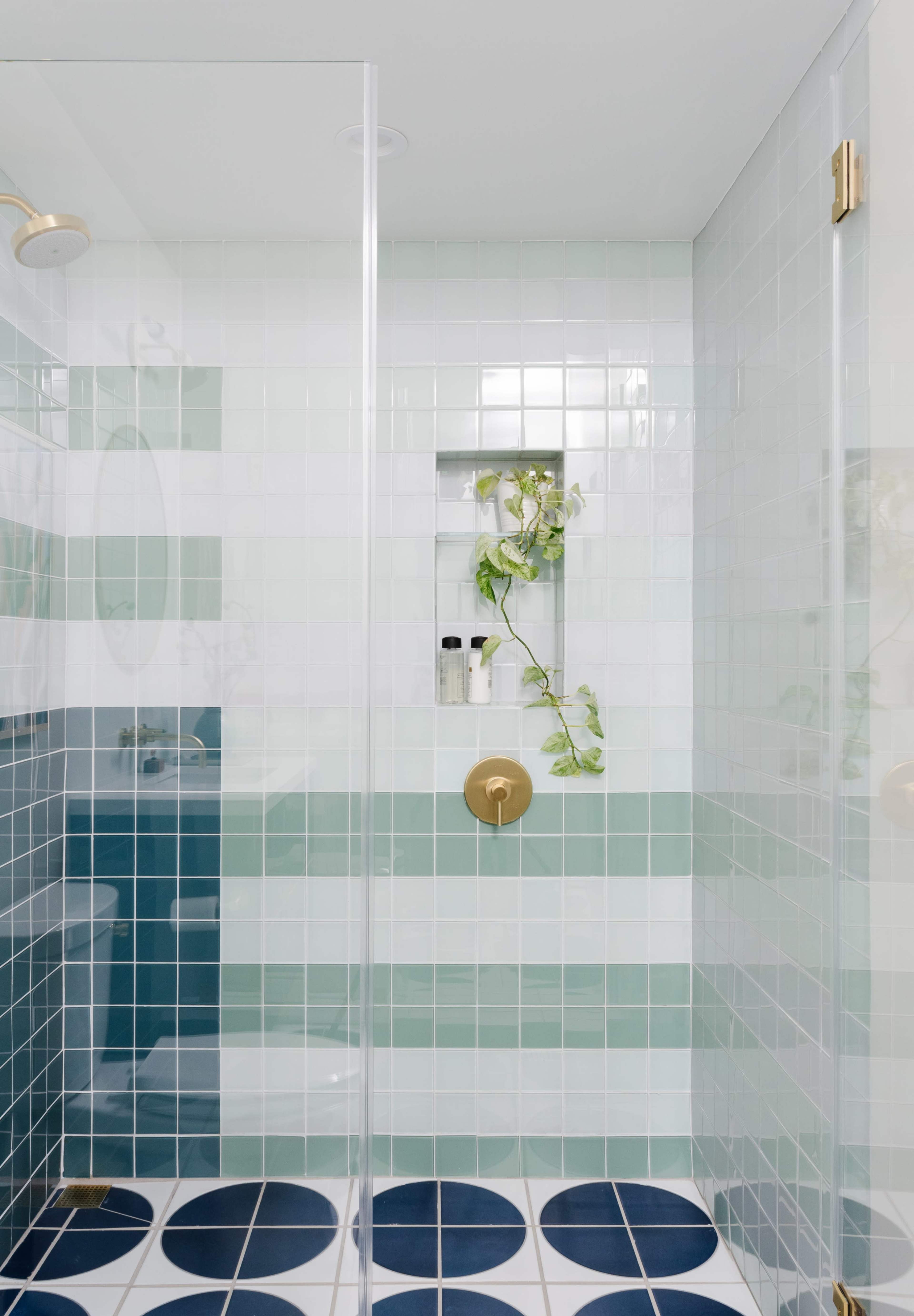 A modern shower with green and blue tiled walls, a glass enclosure, and a niche containing toiletries and a small plant.