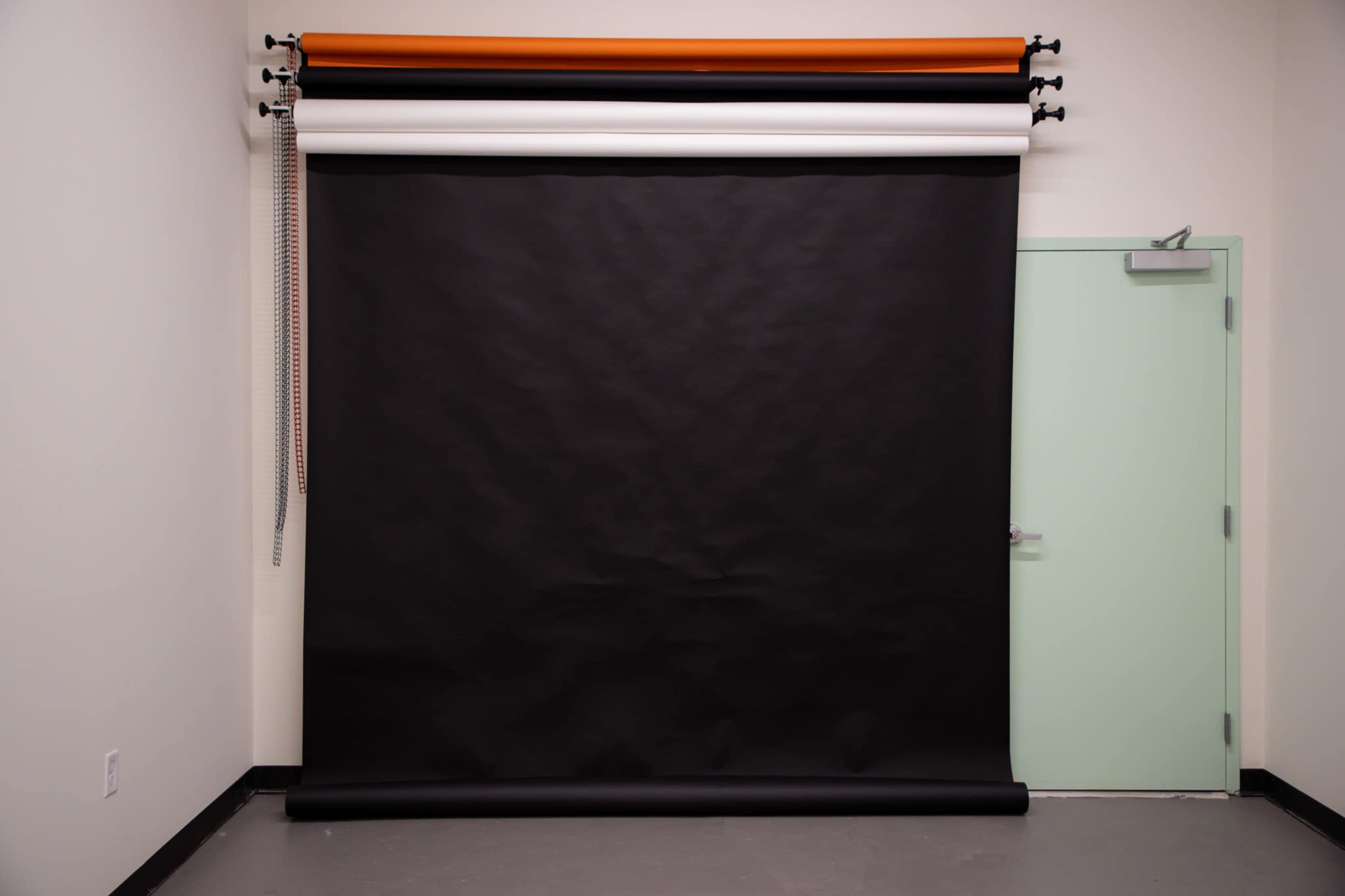 A seamless black backdrop hanging in a studio with orange, white, and black rolls above it and a green door to the right.