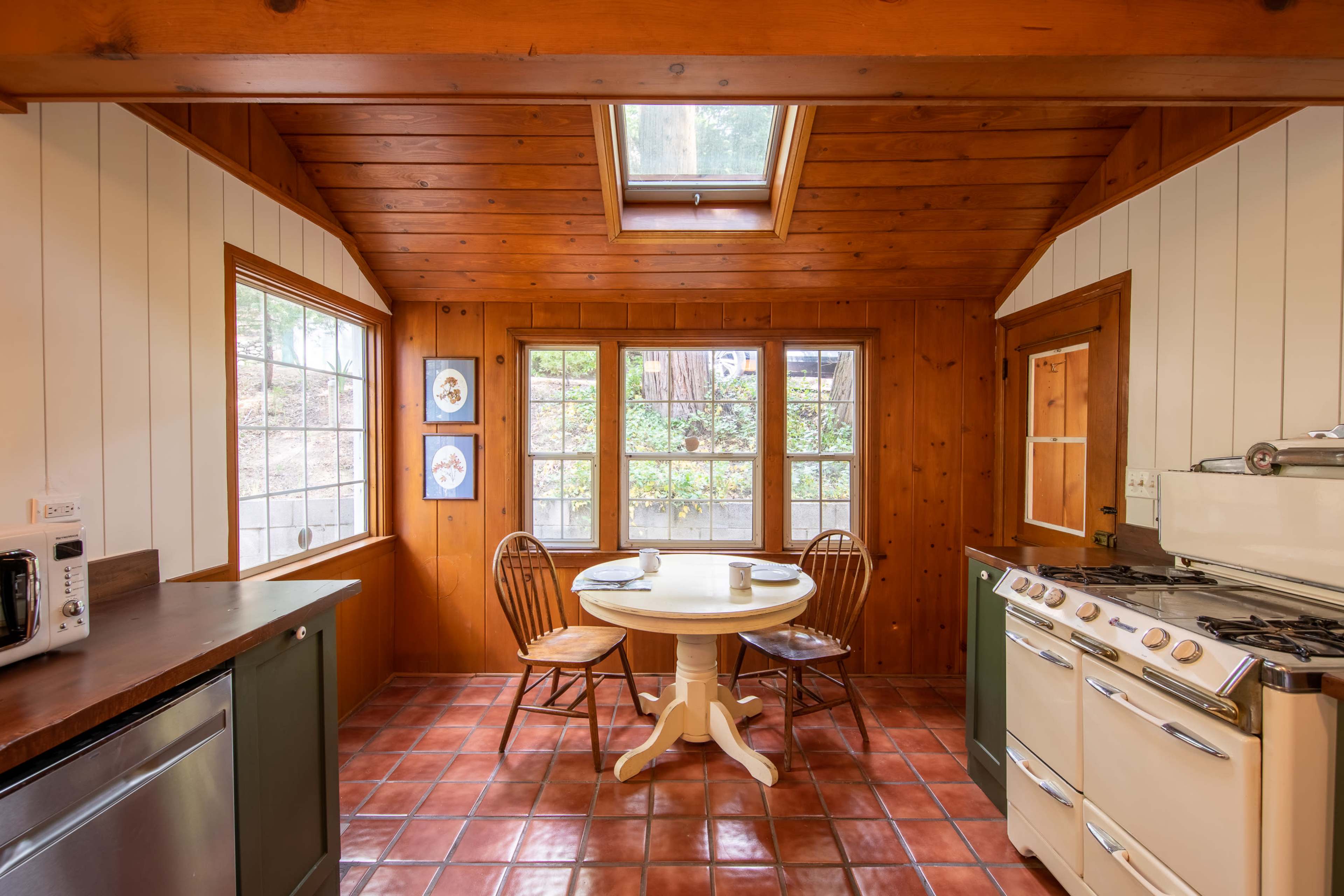 The image shows a cozy kitchen with wooden walls and a round table set for two next to large windows.