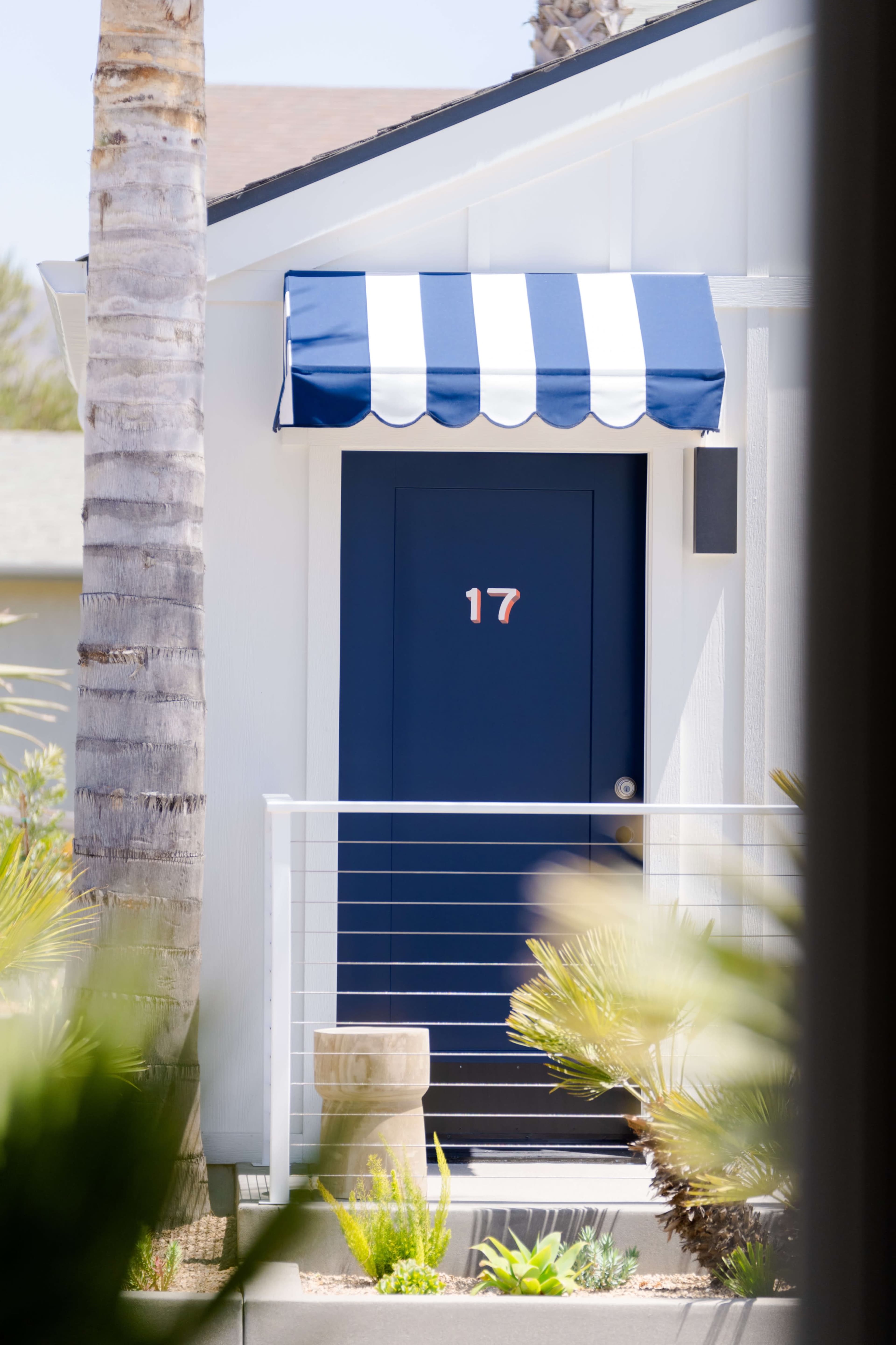 A blue door with the number 17 is located beneath a striped awning, surrounded by palm trees and greenery.