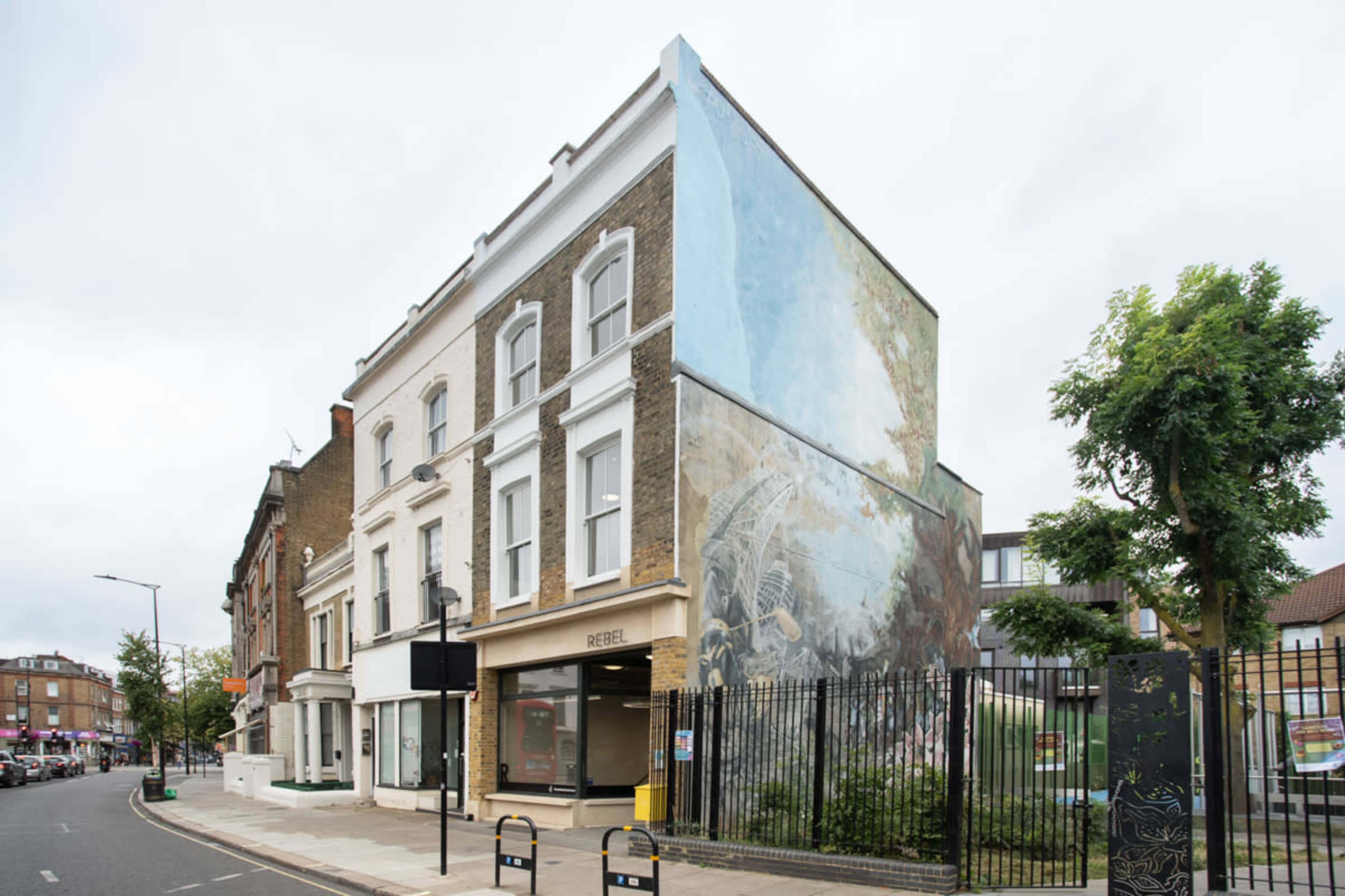 A three-story building with a large mural on its side, situated on a city street lined with trees and parked cars.