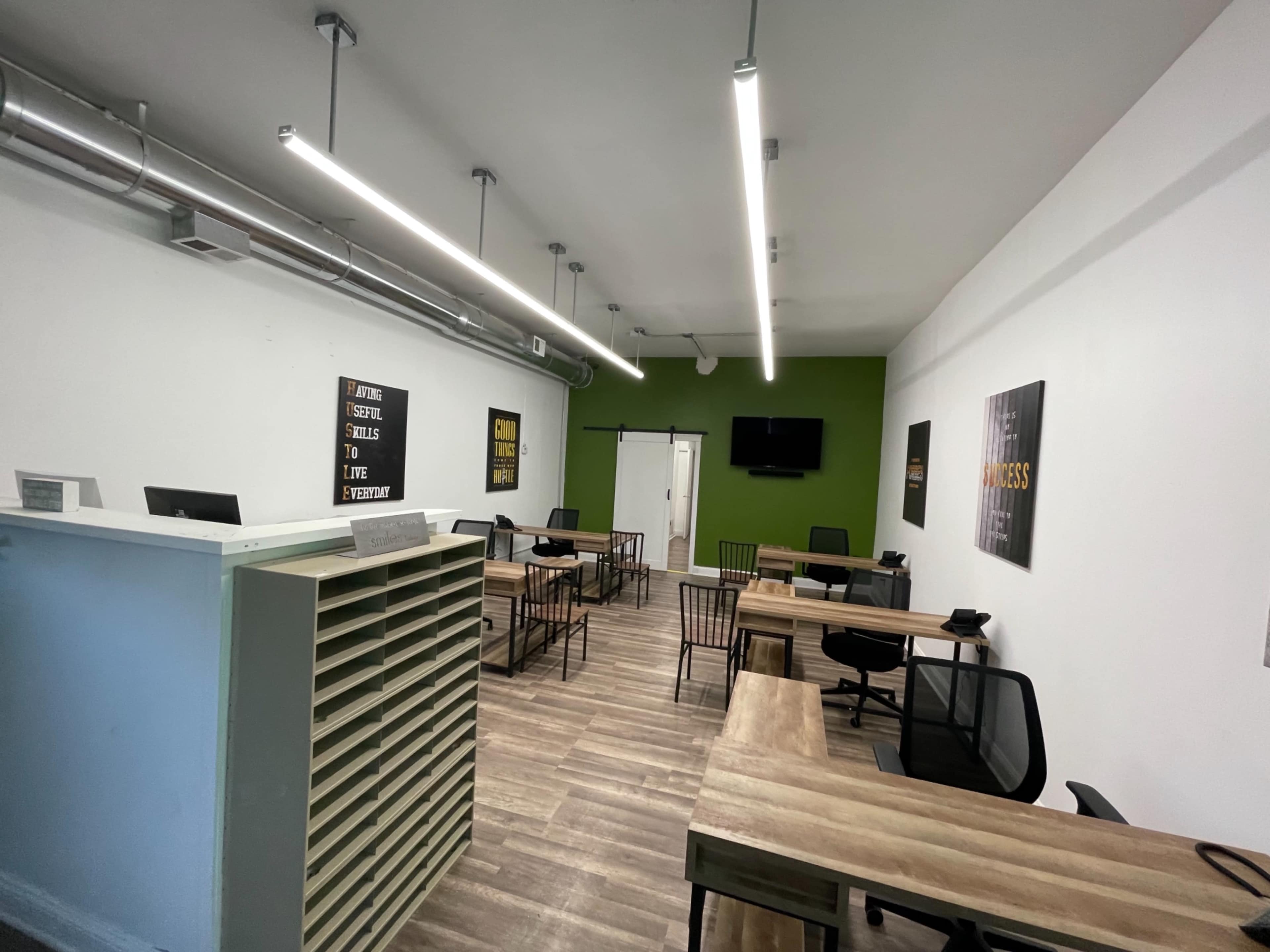 A modern office space with several wooden desks, black chairs, and motivational wall art, featuring a green accent wall.