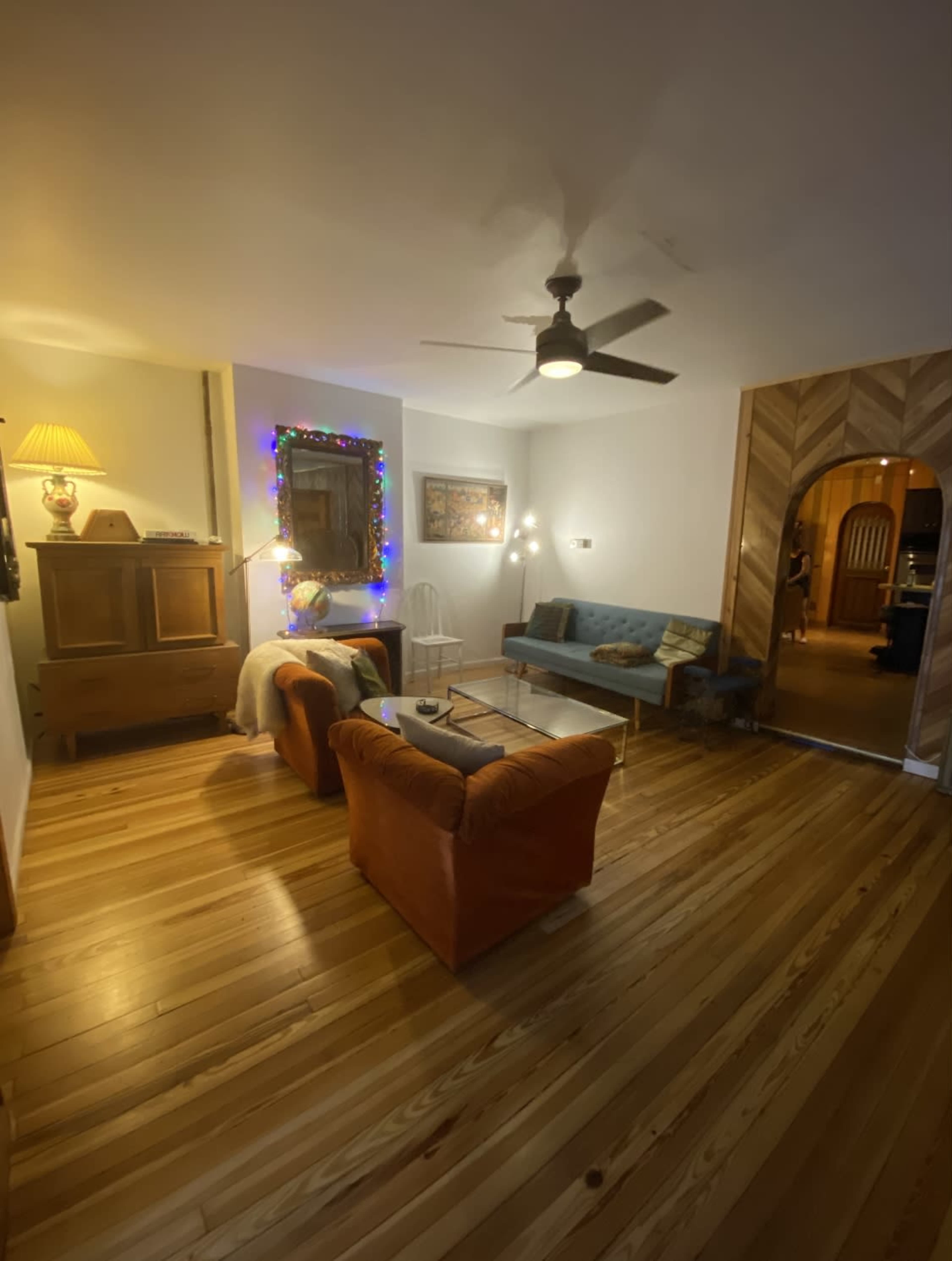 A living room with wooden floors, a blue couch, two orange chairs, and a ceiling fan, illuminated by soft lighting and adorned with decorations.