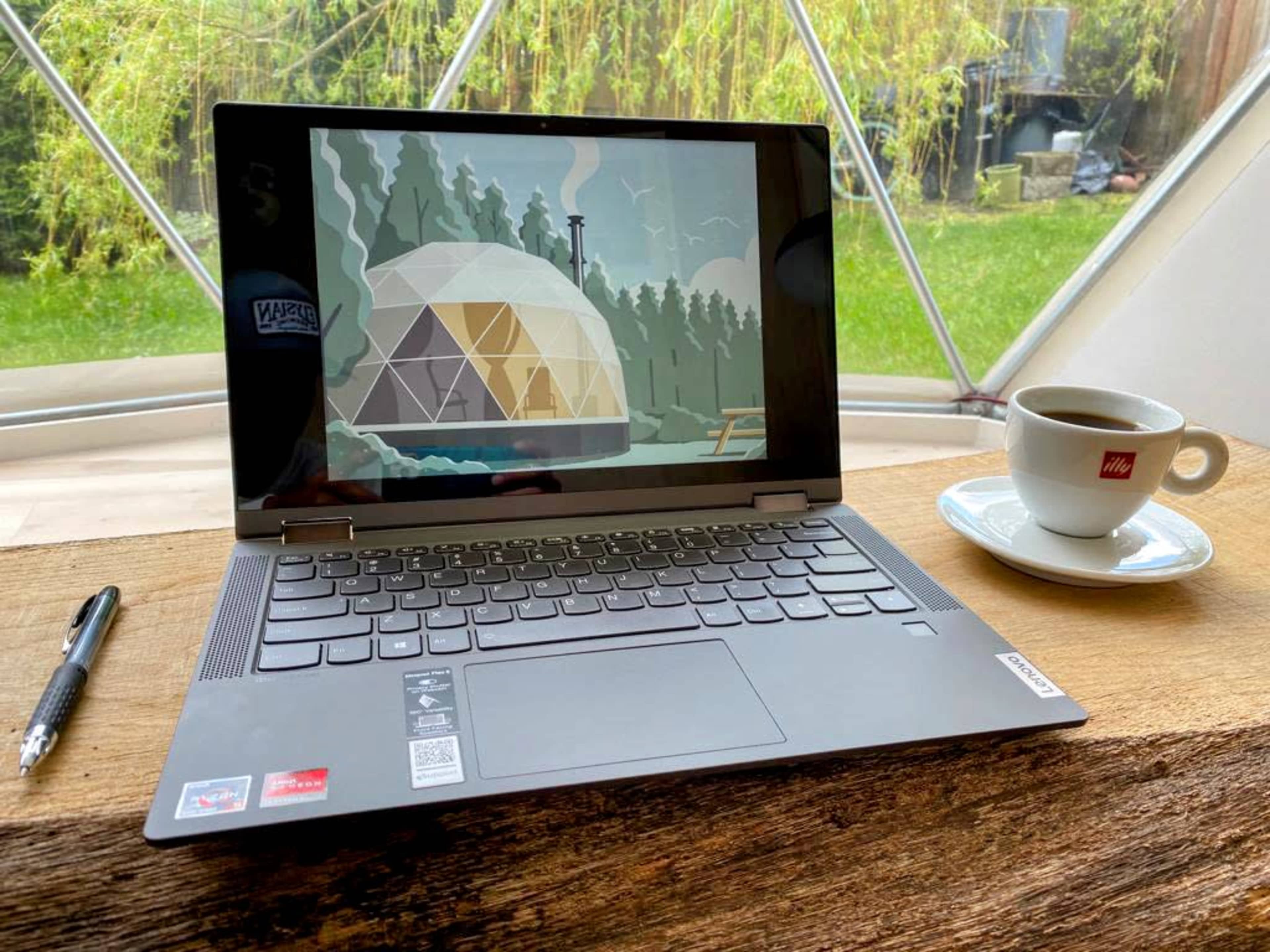A laptop displaying a geometric dome structure is positioned on a wooden table next to a cup of coffee, with a grassy area visible through a large window in the background.