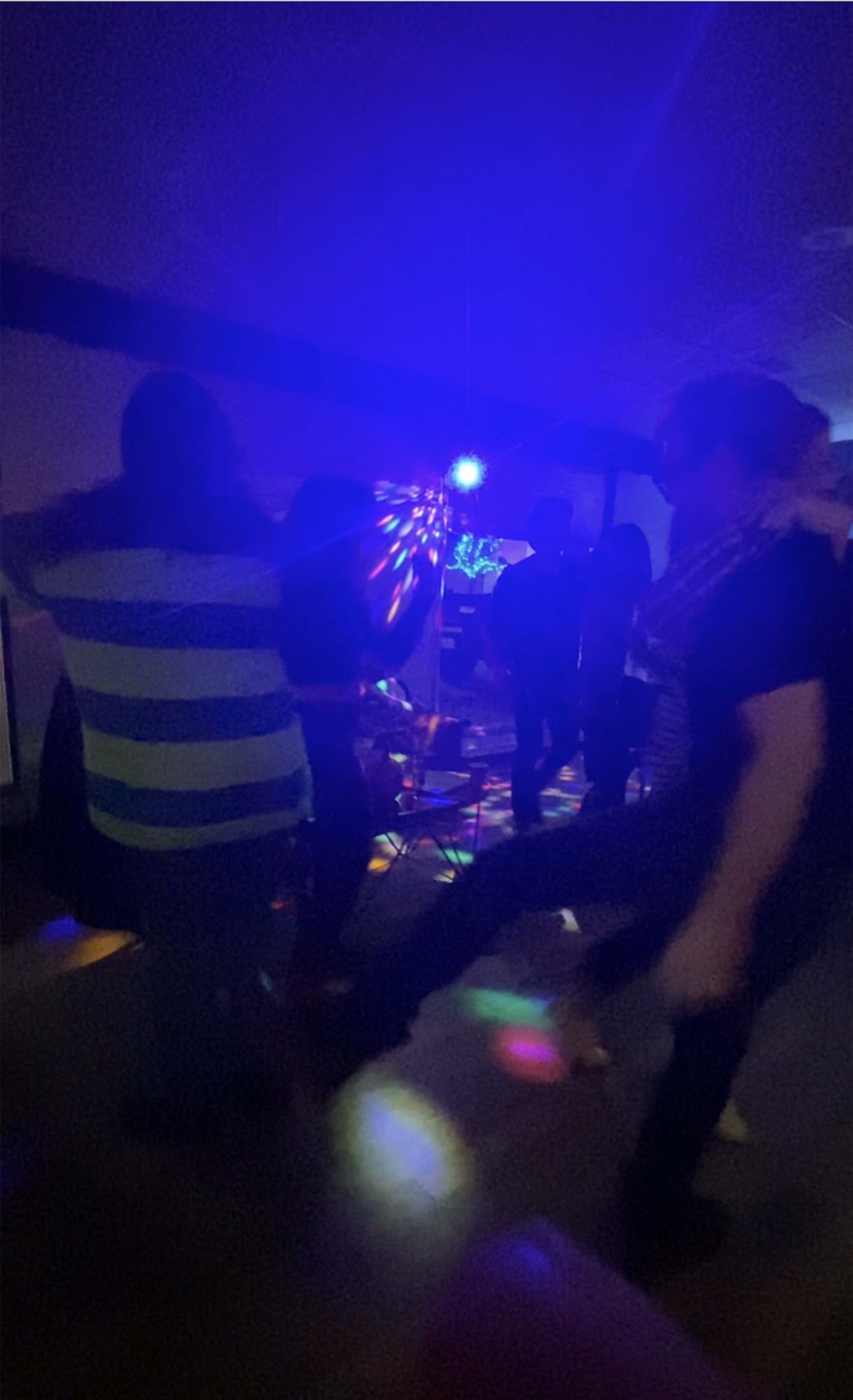 A group of people dances under colorful lights in a dimly lit room.