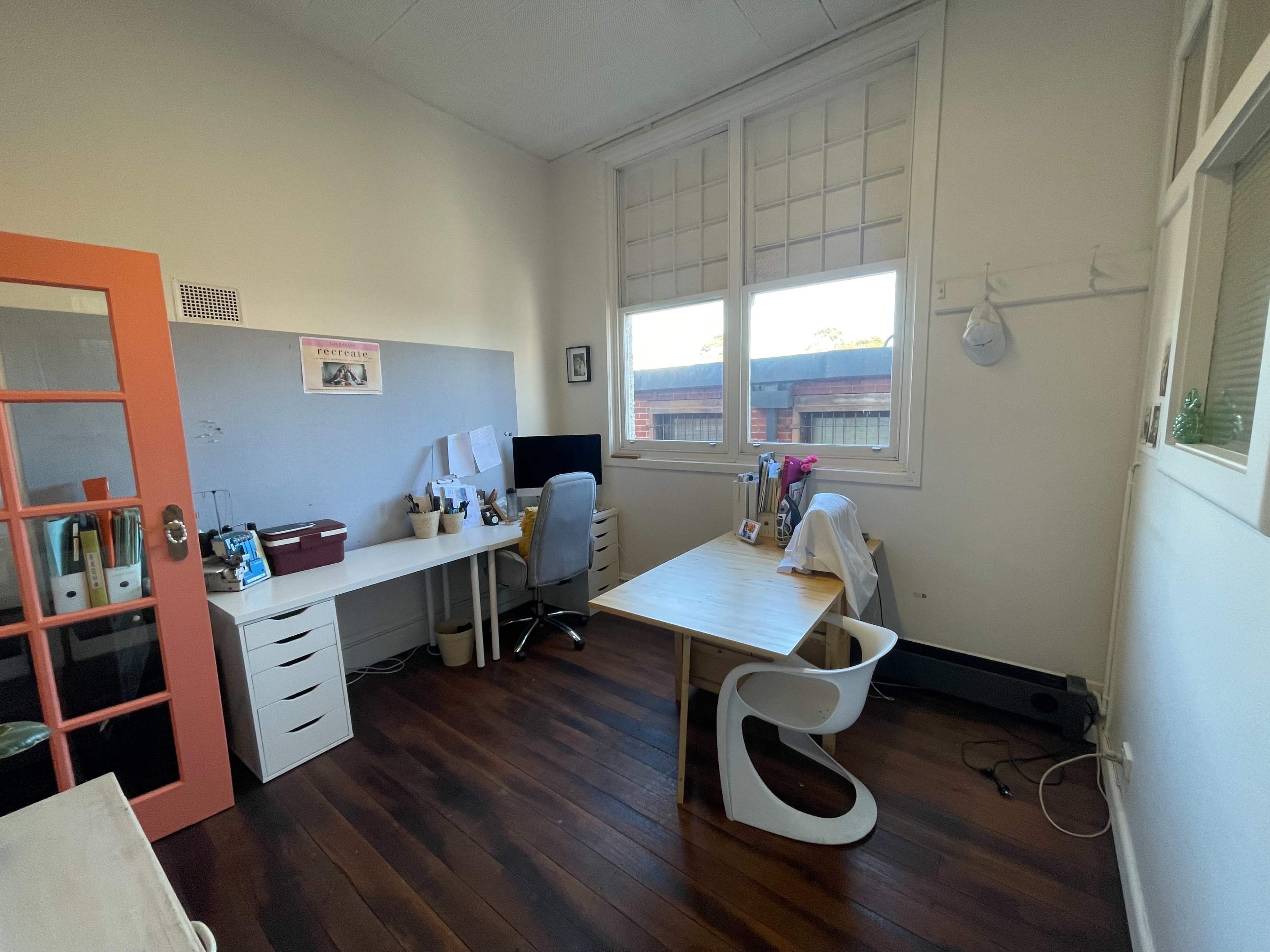 The image shows a small office space featuring a desk with a chair, a computer monitor, and a cabinet, along with large windows allowing natural light to enter.