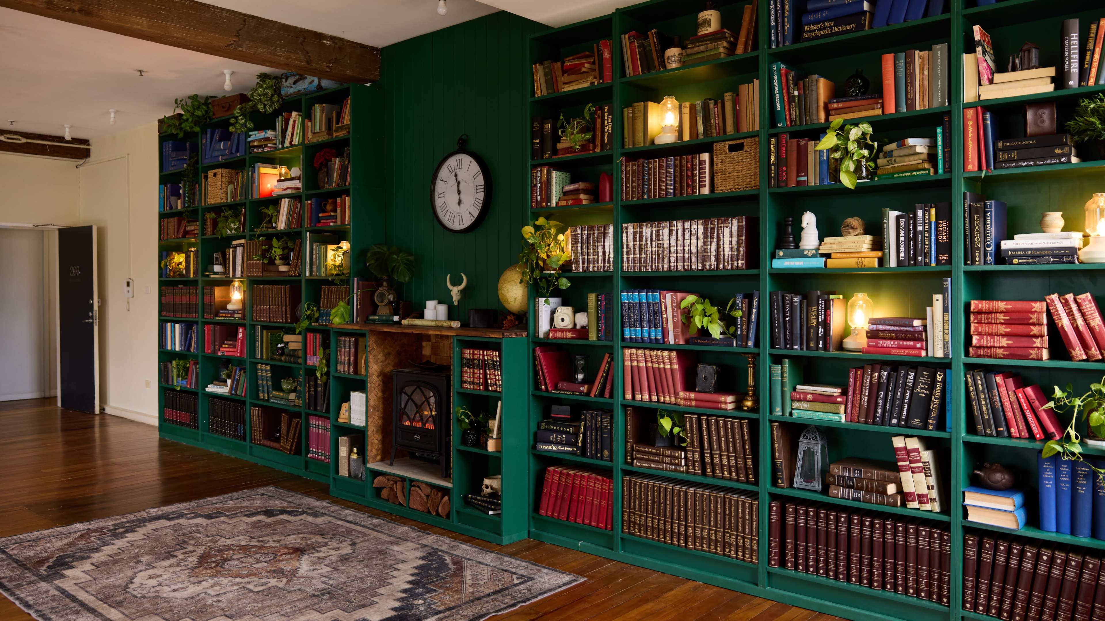 The image features a cozy room with dark green bookshelves filled with books and decorative items, a small fireplace with a mantel, and a patterned rug on the wooden floor.