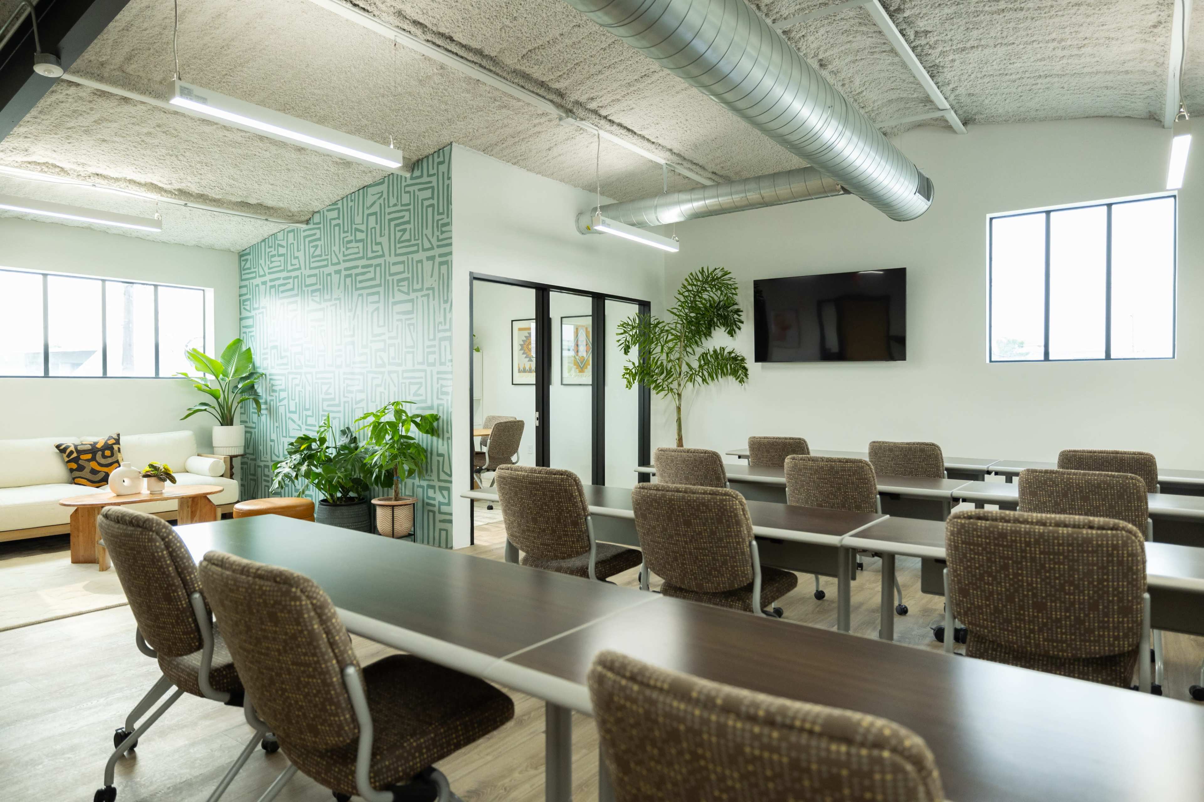 A modern meeting room with multiple tables and chairs arranged in rows, plants in the corners, and a television mounted on the wall.