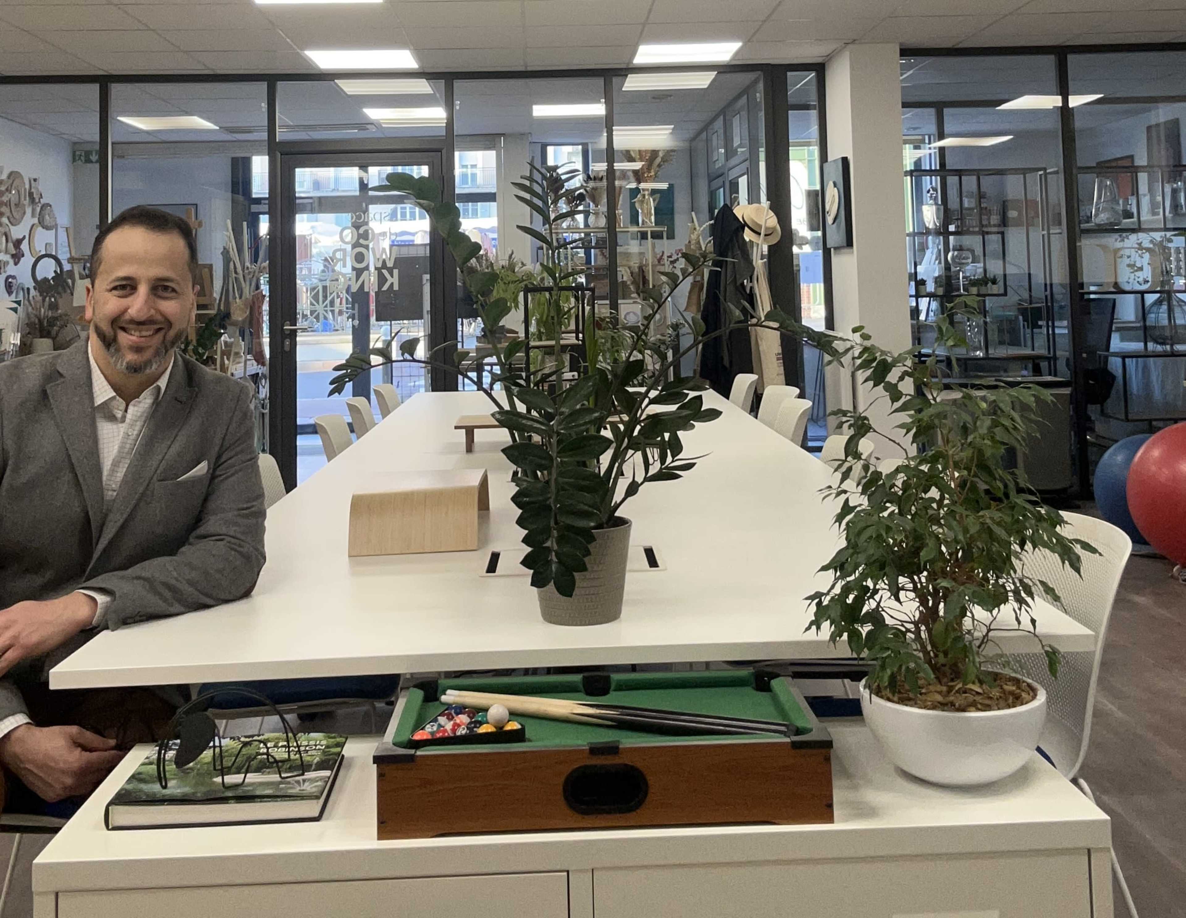 A man in a suit sits beside a large table adorned with plants and a small pool table in a modern office space.