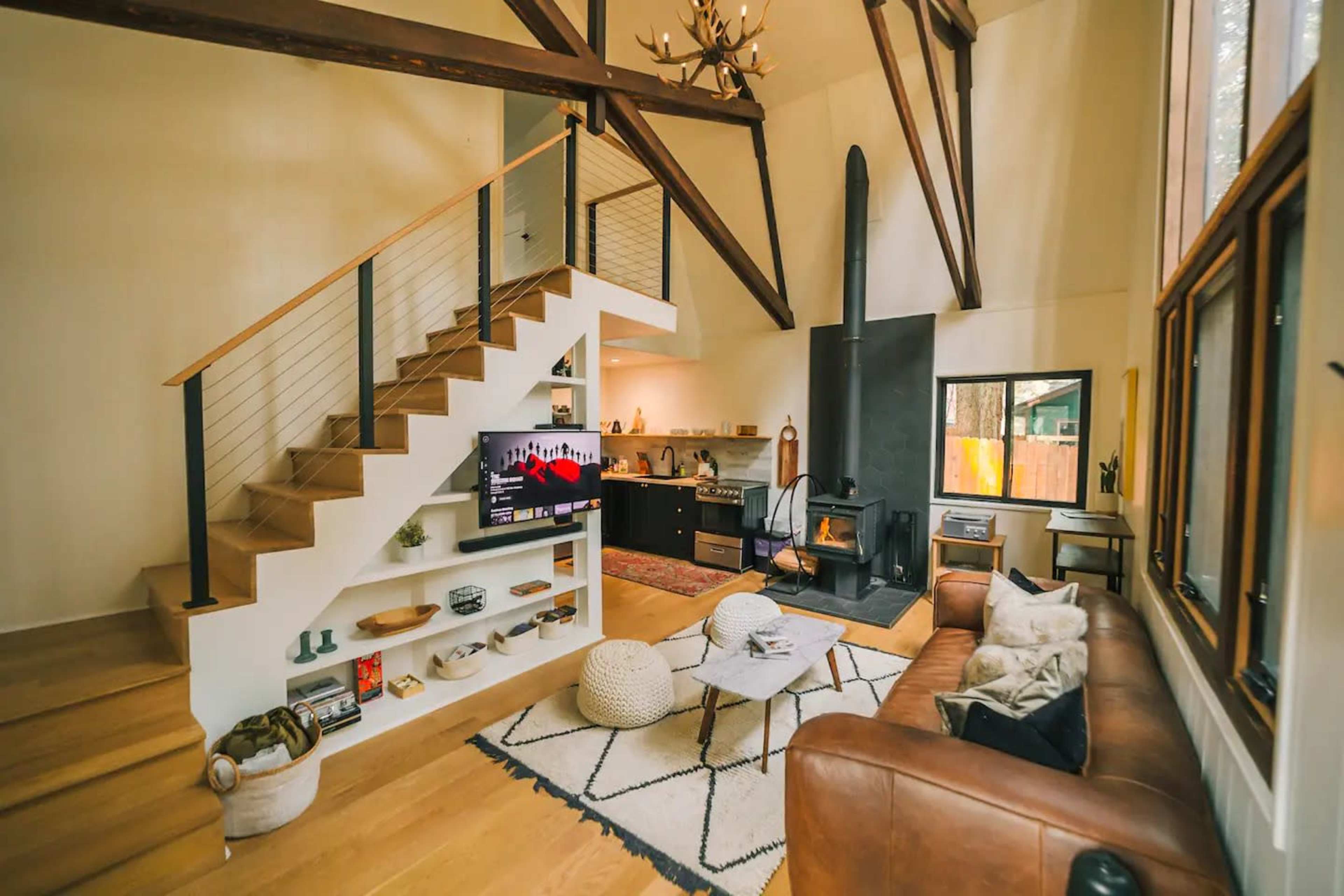 A modern living room with a staircase, a large black fireplace, a TV on a white shelf, and a brown leather couch.