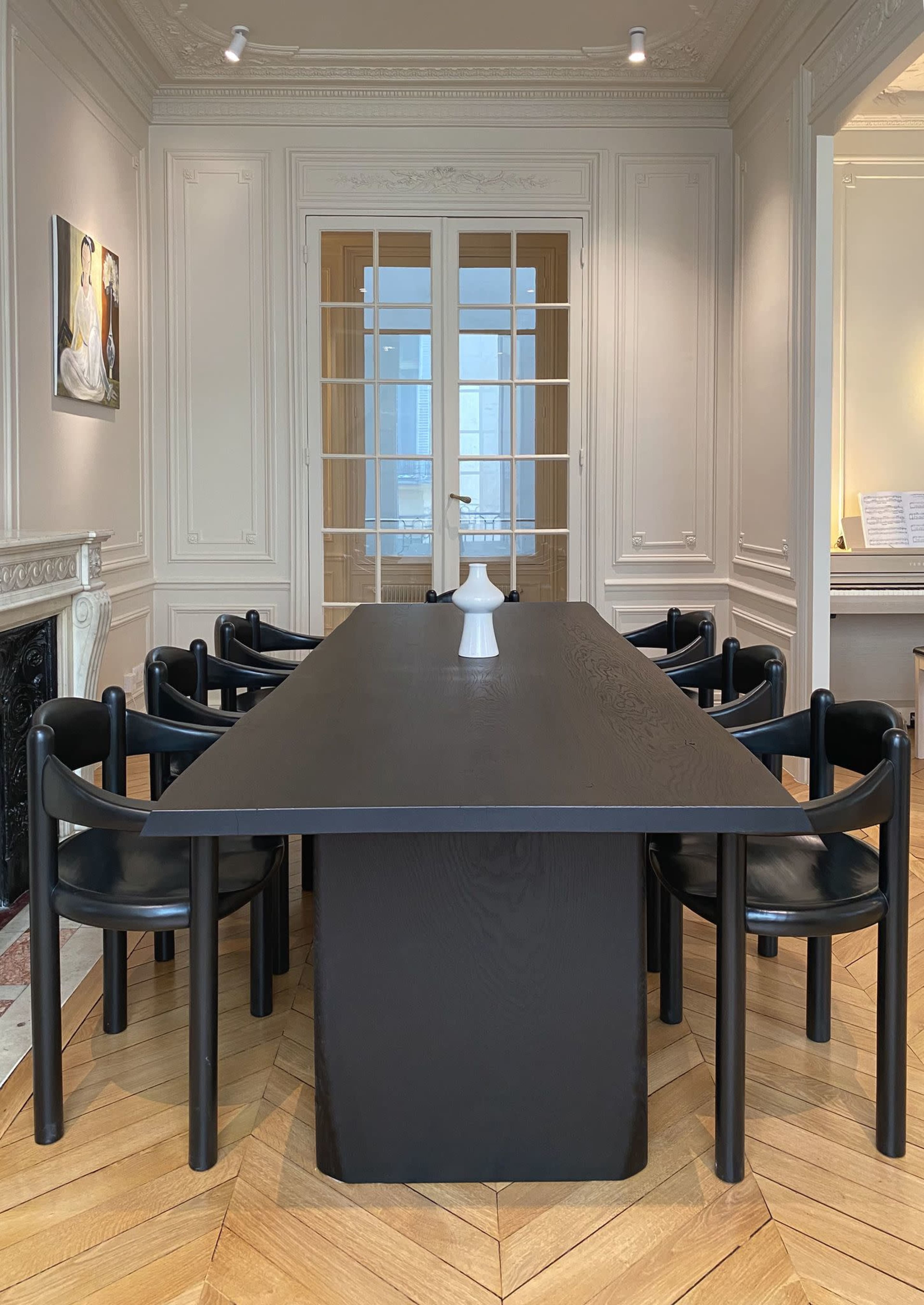 A long black dining table with a white vase is surrounded by six black chairs in a well-lit room with light-colored walls and wooden flooring.